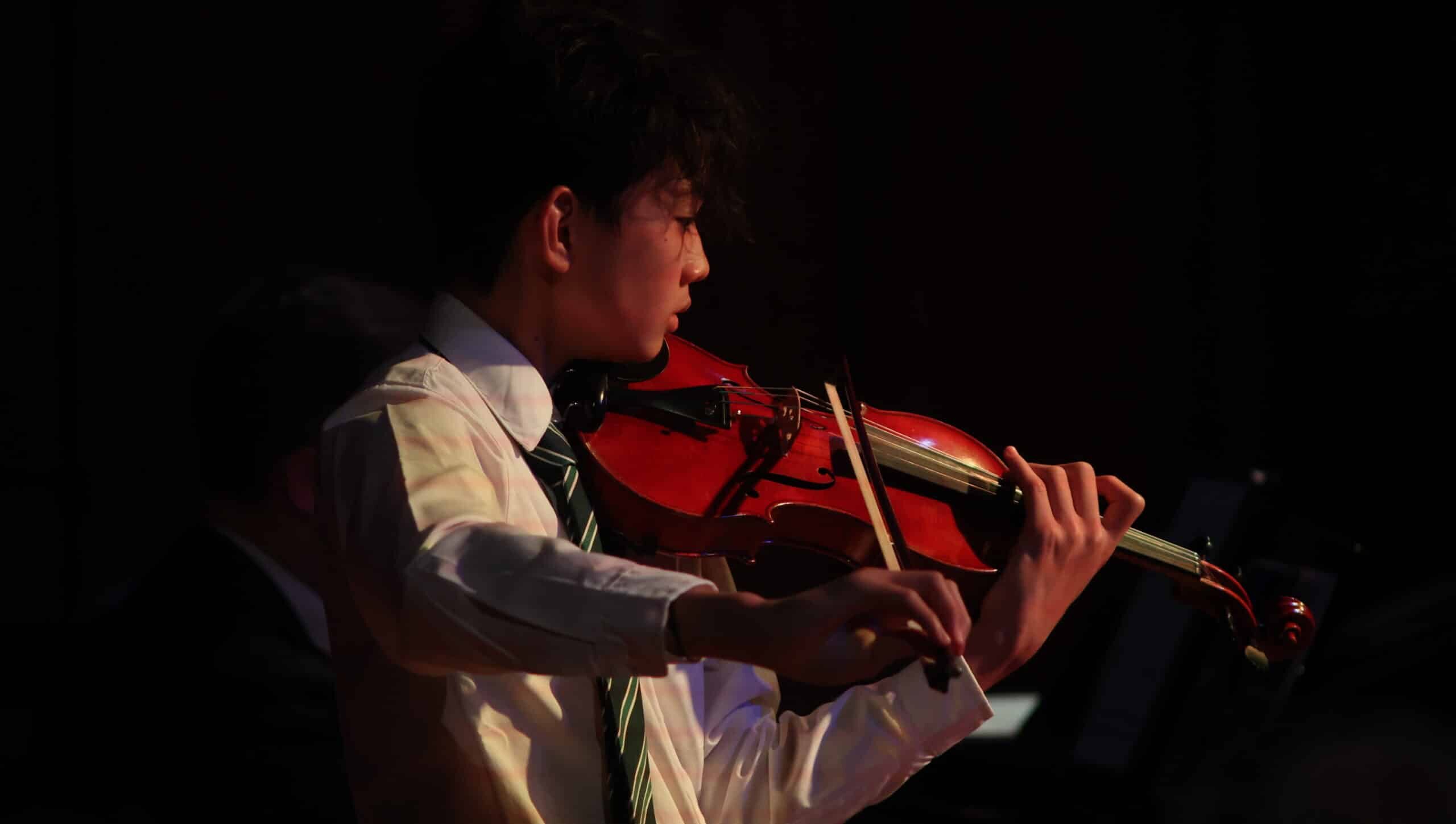 A person in a white shirt and striped tie, perhaps participating in a Specialist Music Programme, plays a violin, focussing intently. The background is dark, and the lighting highlights the musician and the instrument.