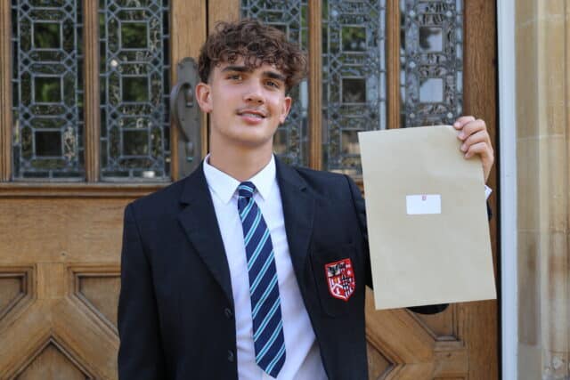 A student in a school uniform stands outside holding up a large brown envelope, smiling with excitement after receiving his GCSE results, with ornate wooden doors and stained glass behind him.