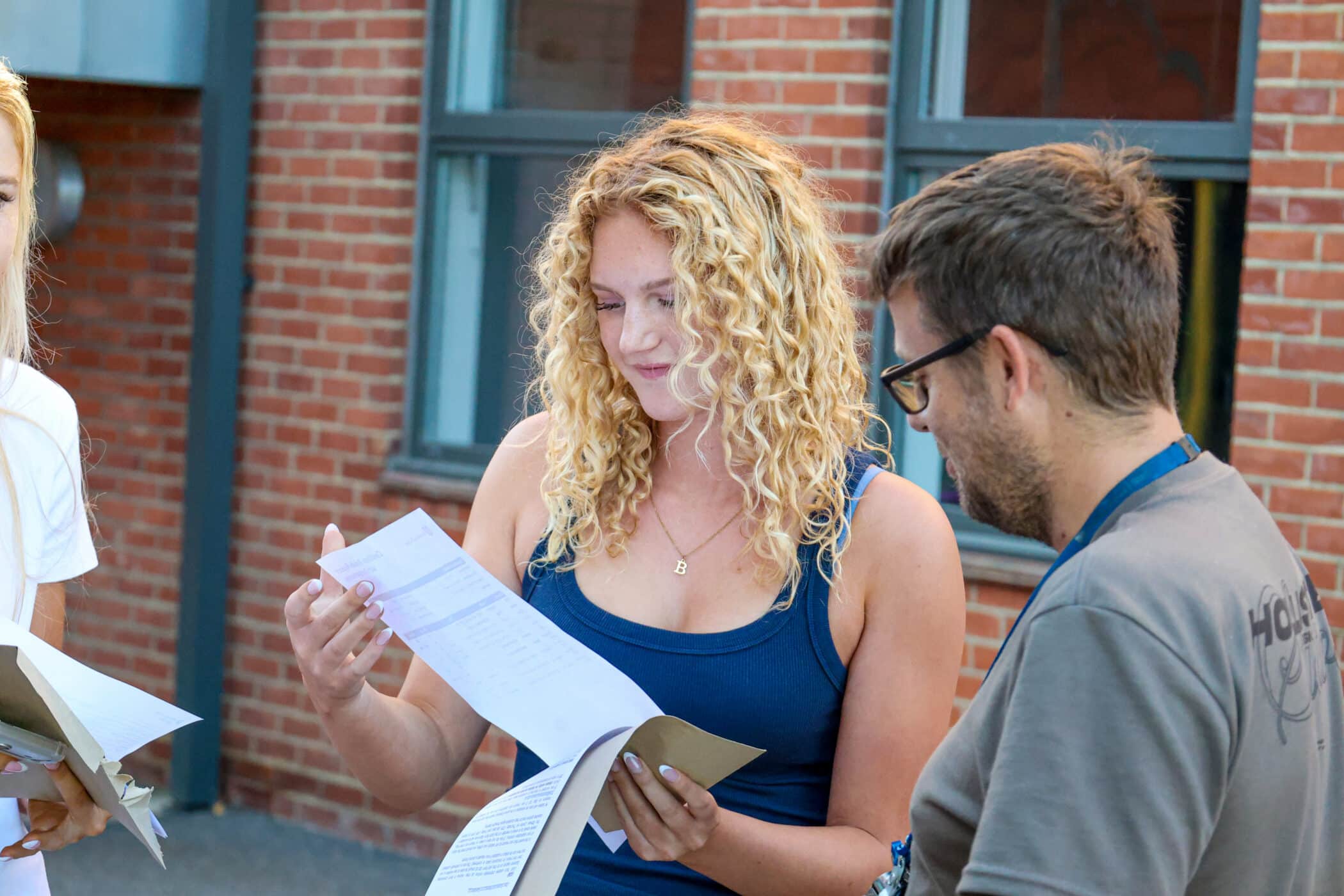 A young woman with curly hair smiles whilst examining a piece of paper, standing next to a man in glasses, with a brick building in the background.