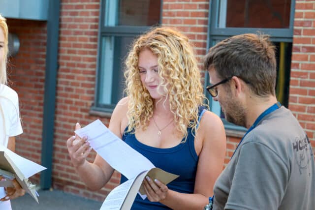 A young woman with curly hair smiles whilst examining a piece of paper, standing next to a man in glasses, with a brick building in the background.
