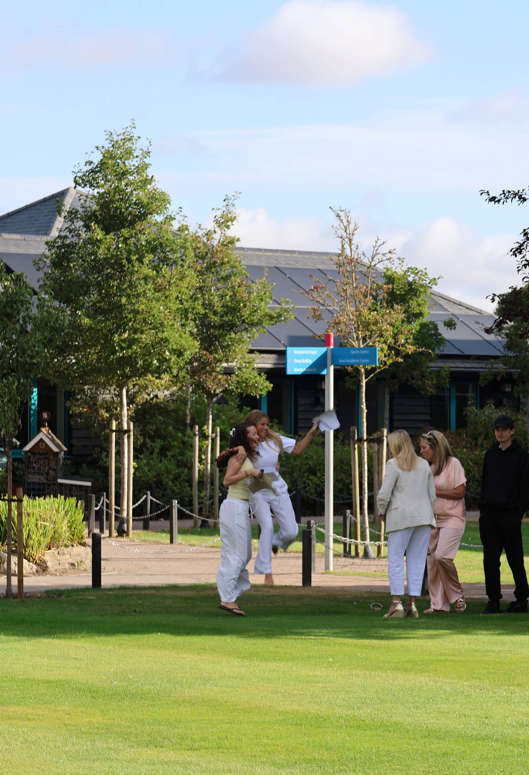 A group of people, mostly dressed in white or light colours, stand and interact on a grassy lawn near a building. Two women are mid-jump and hugging, while others look on. Trees and a blue sky are in the background.