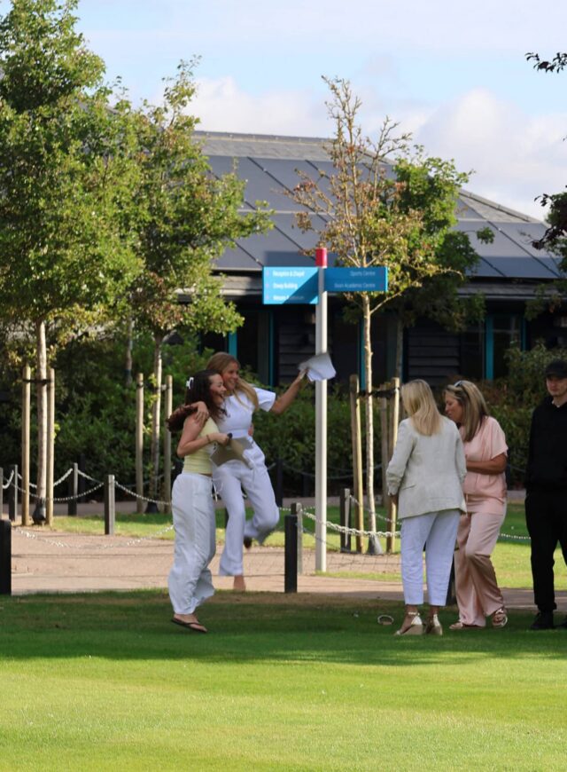A group of people, mostly dressed in white or light colours, stand and interact on a grassy lawn near a building. Two women are mid-jump and hugging, while others look on. Trees and a blue sky are in the background.