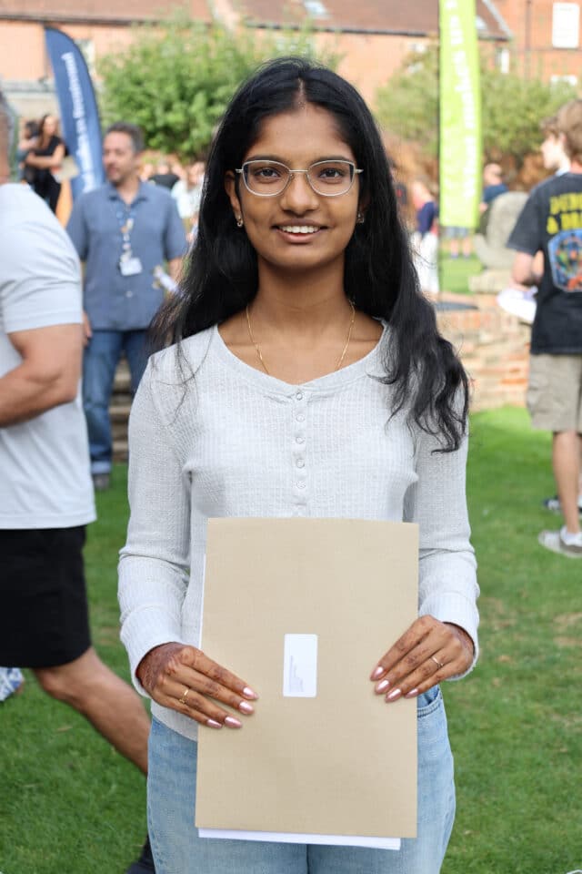 A young woman with glasses stands outdoors, smiling and holding a brown envelope. People and banners are visible in the background at what appears to be an event or gathering.