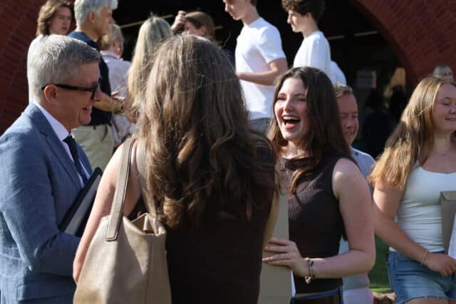 A group of people stand outdoors, smiling and talking after finishing their GCSE exams. One young woman in the foreground laughs, holding a bag. Others are mingling in the background near a red brick archway.