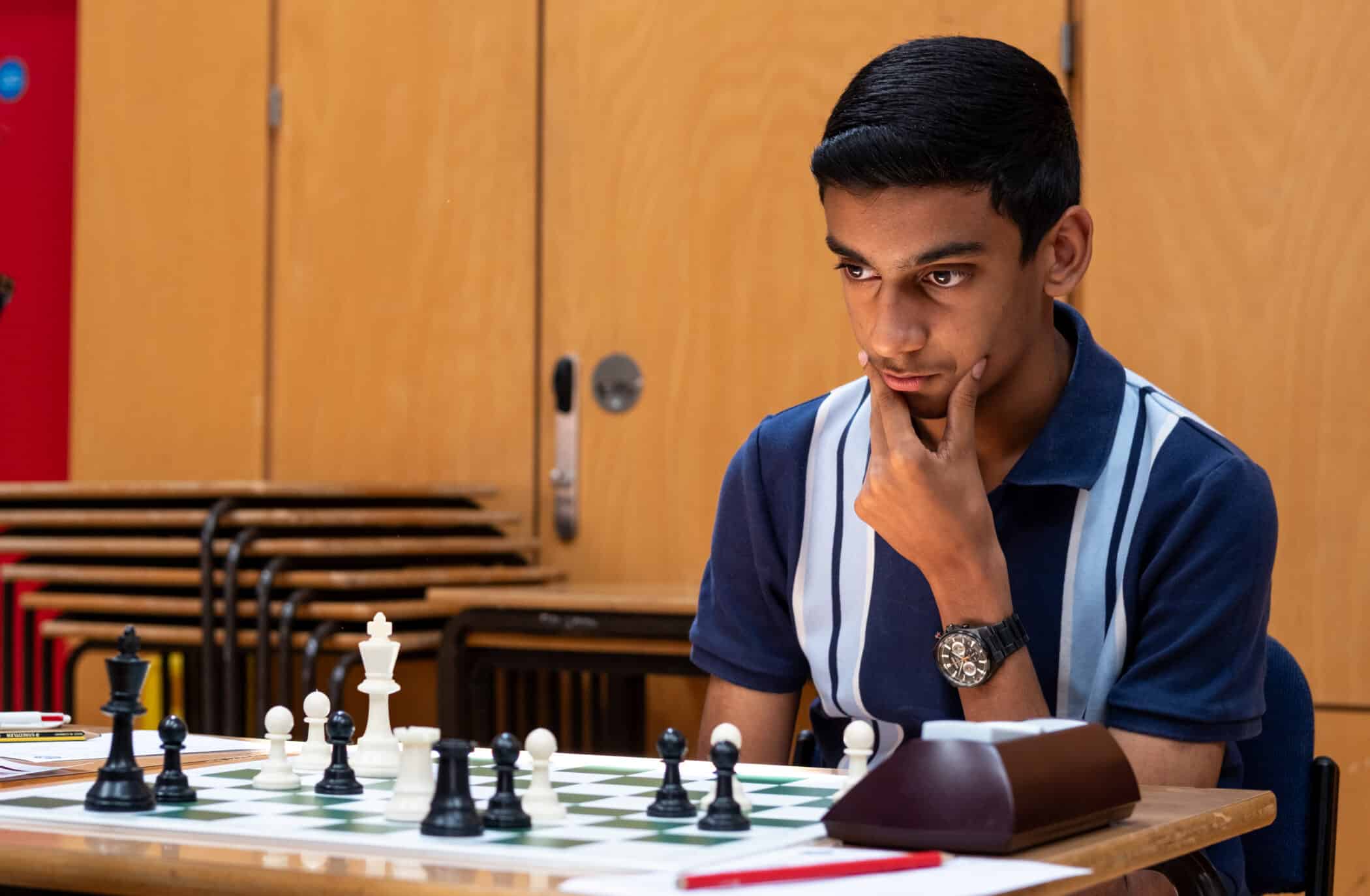 A young person intently studies a chessboard during a game, with their hand on their chin and chess pieces arranged in play. Tables and chairs are in the background.