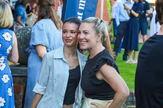Two women stand close together and smile for a photo at an outdoor event. Other people are gathered and mingling in the background. Both women look happy and casually dressed.