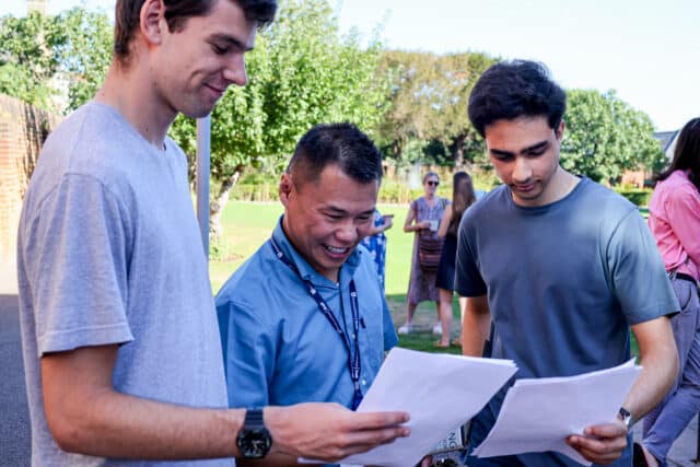 Three people stand outdoors, smiling and holding papers, appearing to discuss results. Other people and green trees are visible in the background under a blue sky.