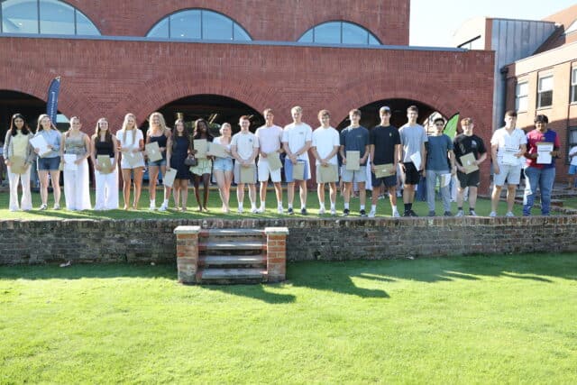 A group of young people stands in a row on a low brick stage outdoors, each holding certificates, with a brick building and green lawn in the background.