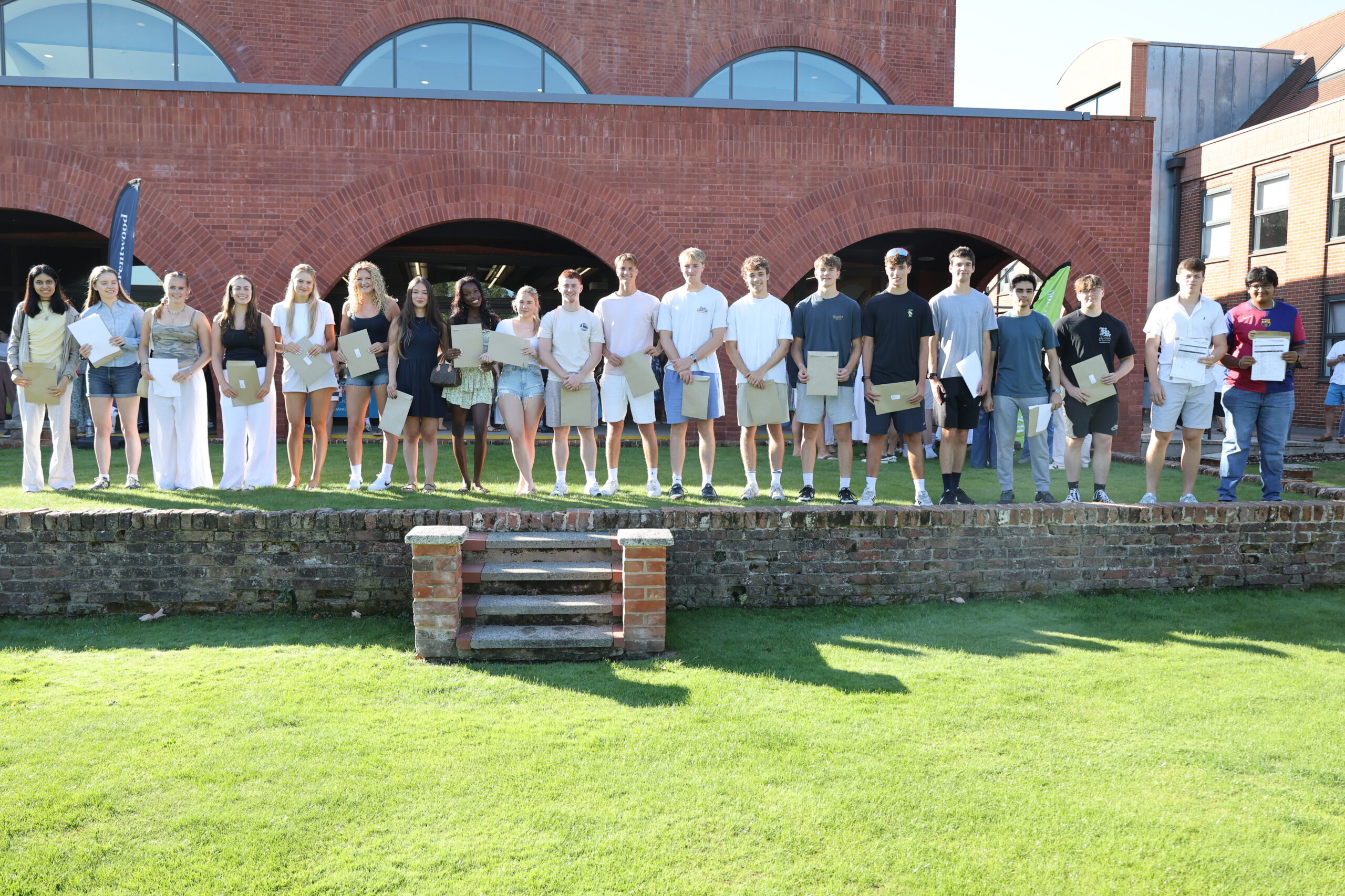 A group of young people stands in a row on a low brick stage outdoors, each holding certificates, with a brick building and green lawn in the background.