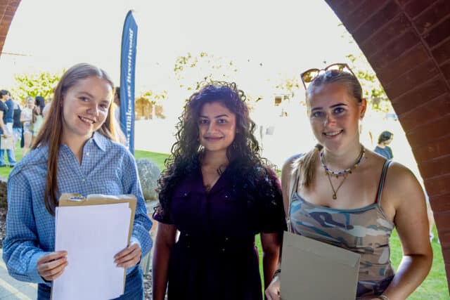Three young women stand under a brick archway, smiling and holding envelopes or papers. Sunlight brightens the background, and other people can be seen on the grass outside.