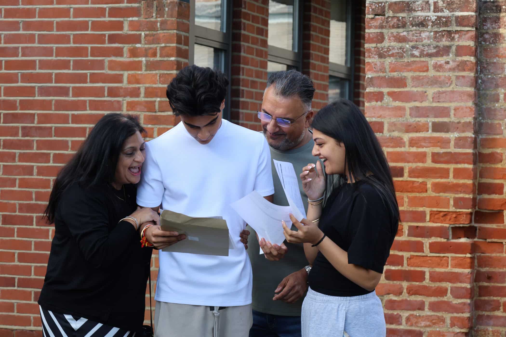 A young man reads a paper whilst standing with three smiling people, possibly his family, outside a brick building. Everyone looks happy and excited as they look at the GCSE results together.