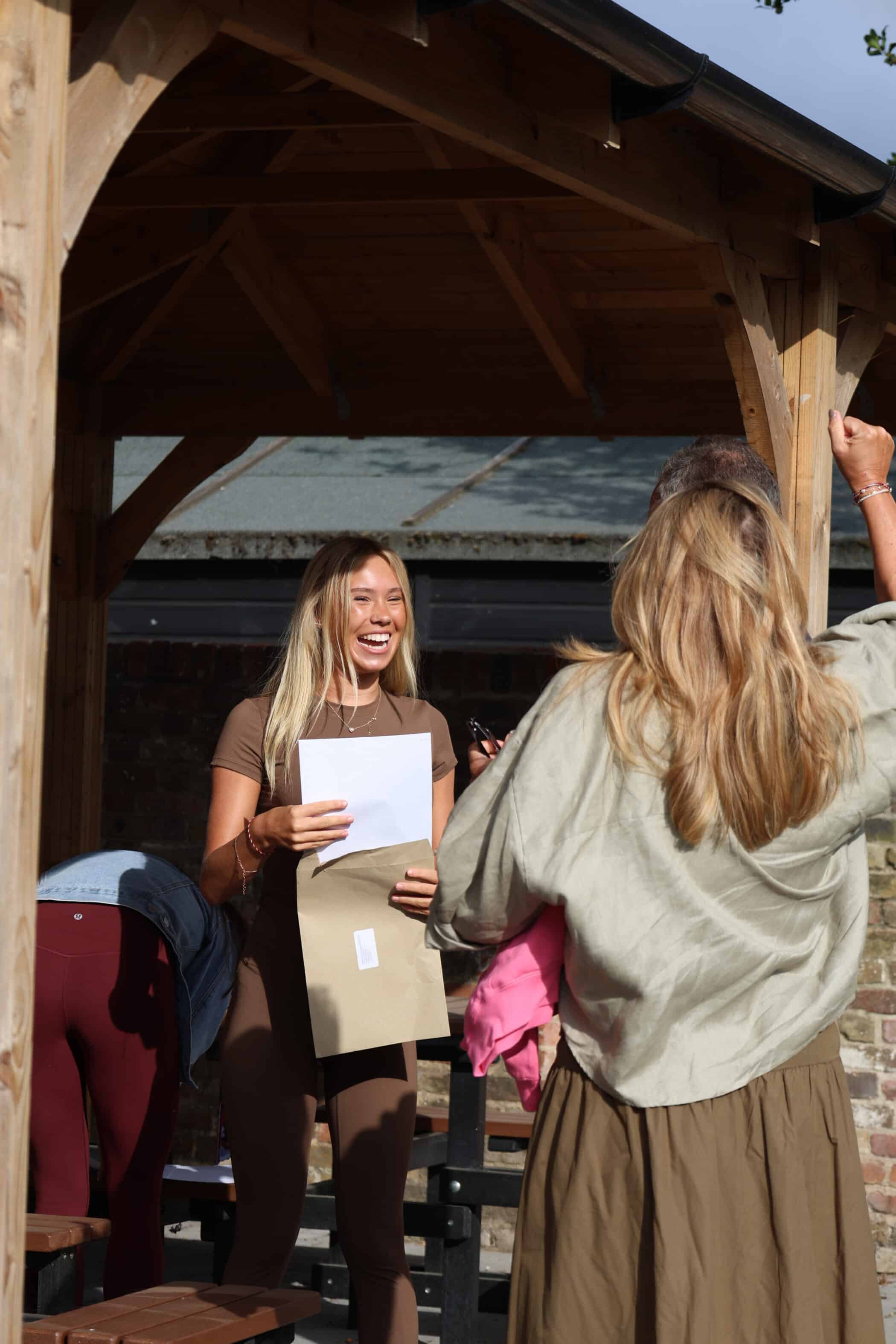 A young woman stands under a wooden shelter, smiling and holding a piece of paper and an envelope containing her GCSE results, whilst another person with long hair faces her. It appears to be a celebratory or happy moment outdoors.