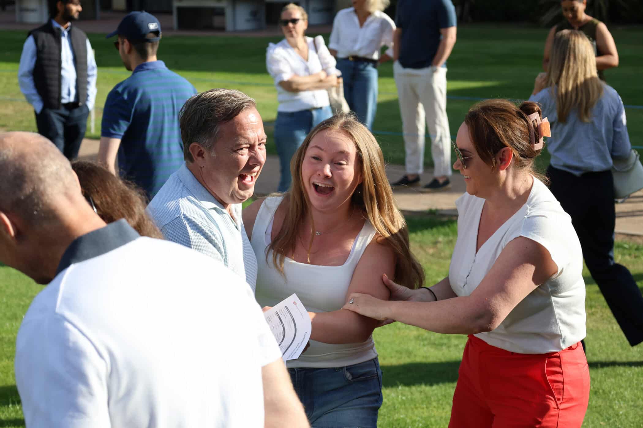 A group of people outdoors laugh and smile together on a sunny day, with two women and a man in the foreground holding GCSE papers and appearing joyful. Other people are in the background on the grass.