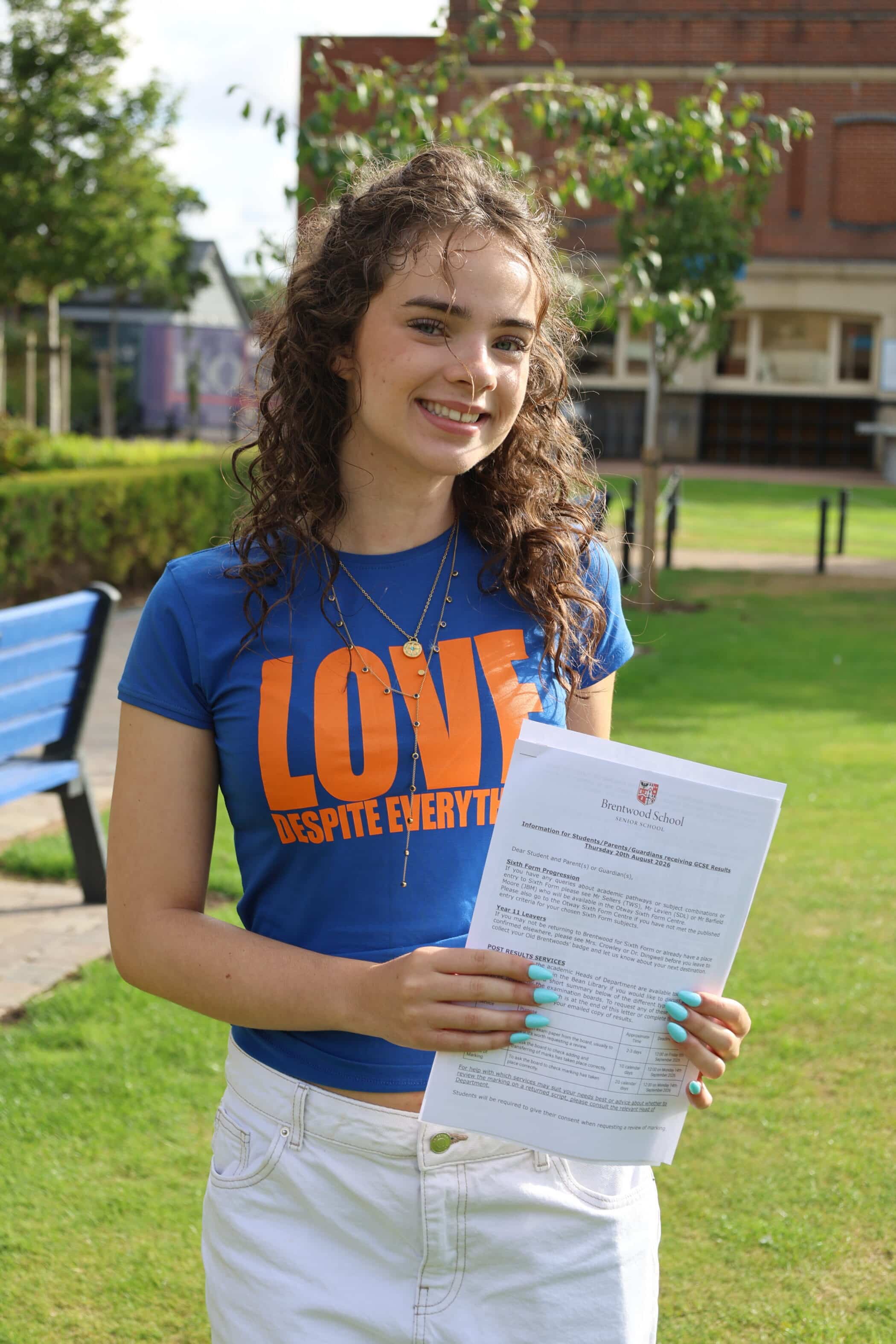 A young woman with curly hair smiles, holding her GCSE document whilst standing outside on a sunny day. She wears a blue shirt with LOVE DESPITE EVERYTHING written in orange letters.