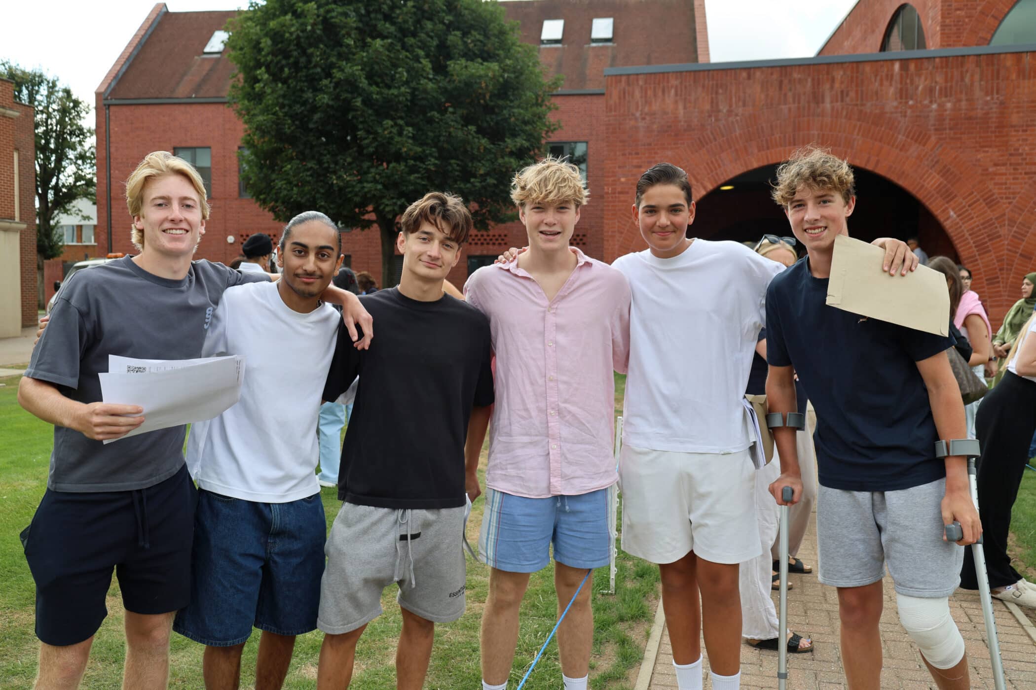 Six teenage boys stand outside smiling arm-in-arm, with one holding GCSE papers and another on crutches. Red brick buildings and a tree are in the background. They appear happy and casually dressed.