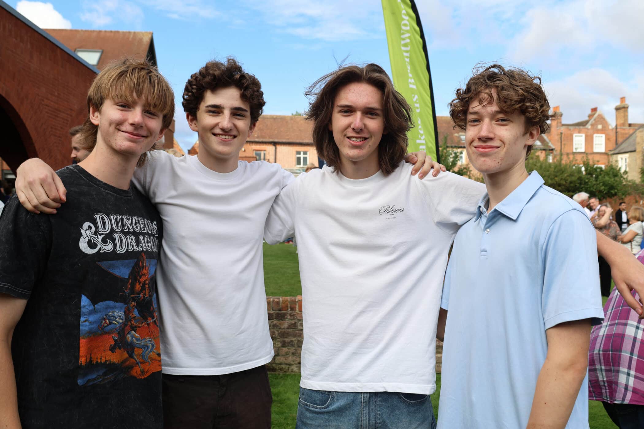 Four young men stand outdoors, smiling with arms round each other after their GCSE results. They are casually dressed, and there are buildings, grass, and a green banner in the background under a partly cloudy sky.