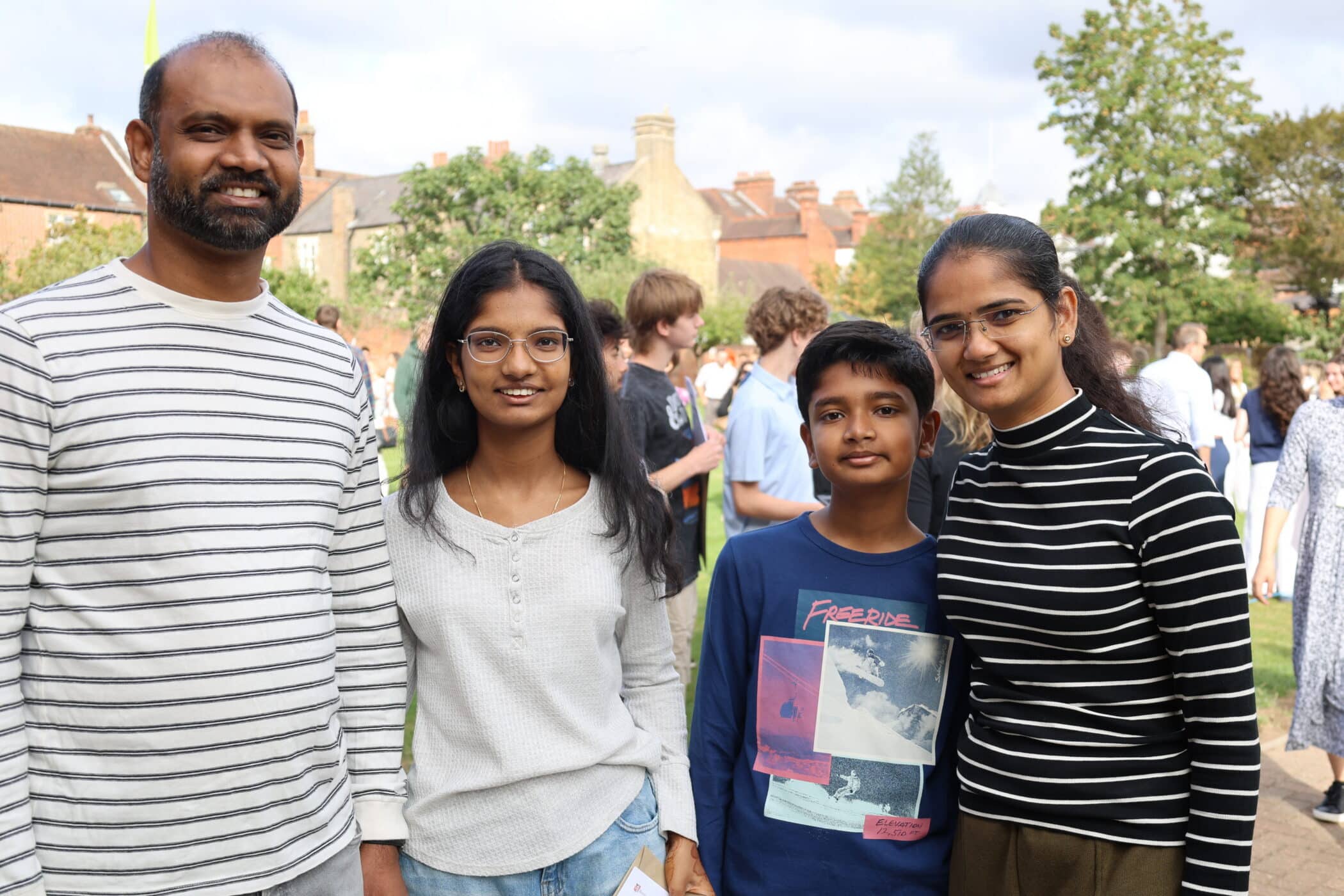 A family of four stands outside, smiling at the camera, perhaps celebrating a recent GCSE achievement. There are people and trees in the background.