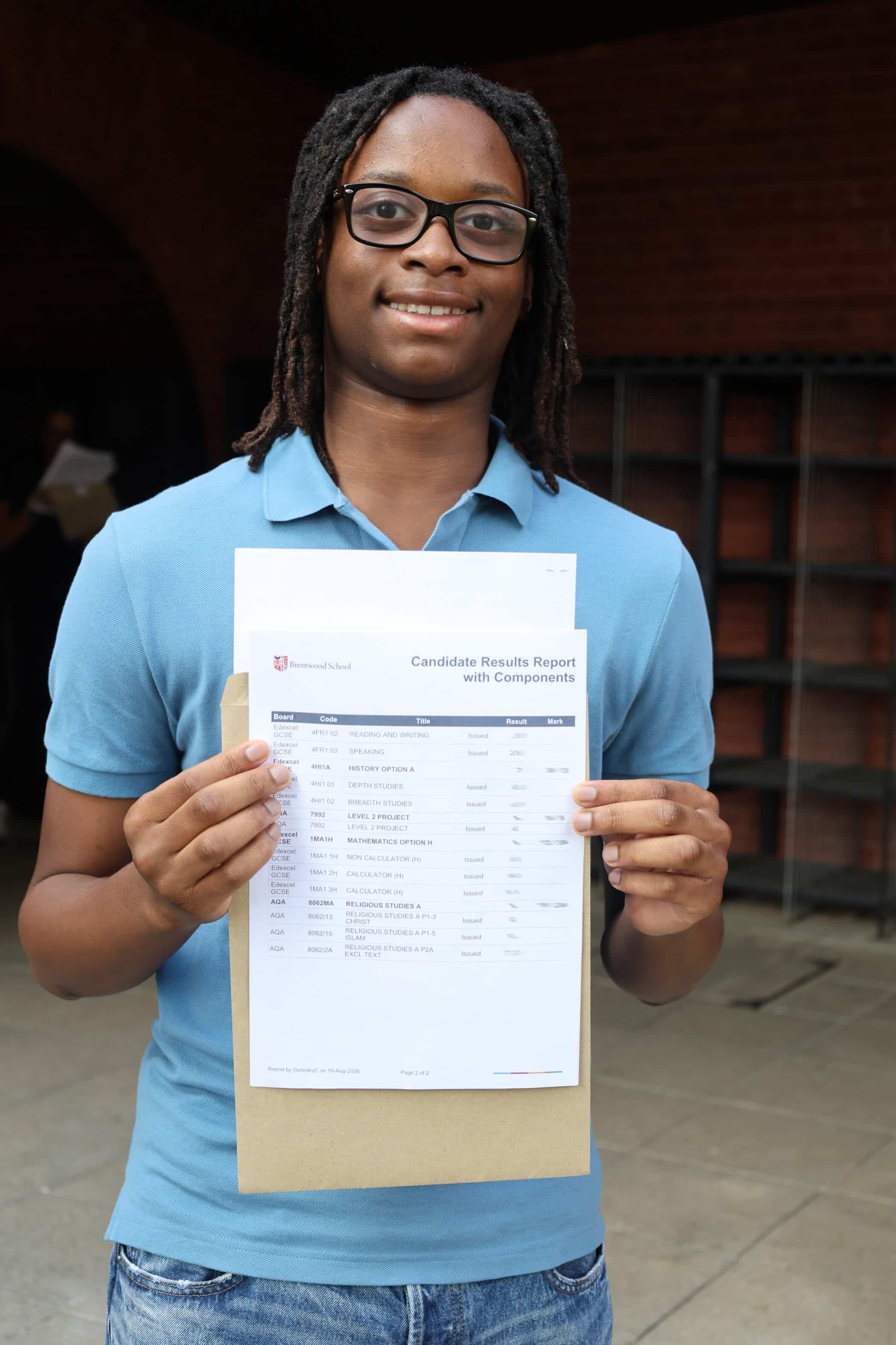 A person wearing glasses and a blue polo shirt smiles whilst holding up a printed GCSE results report outdoors.