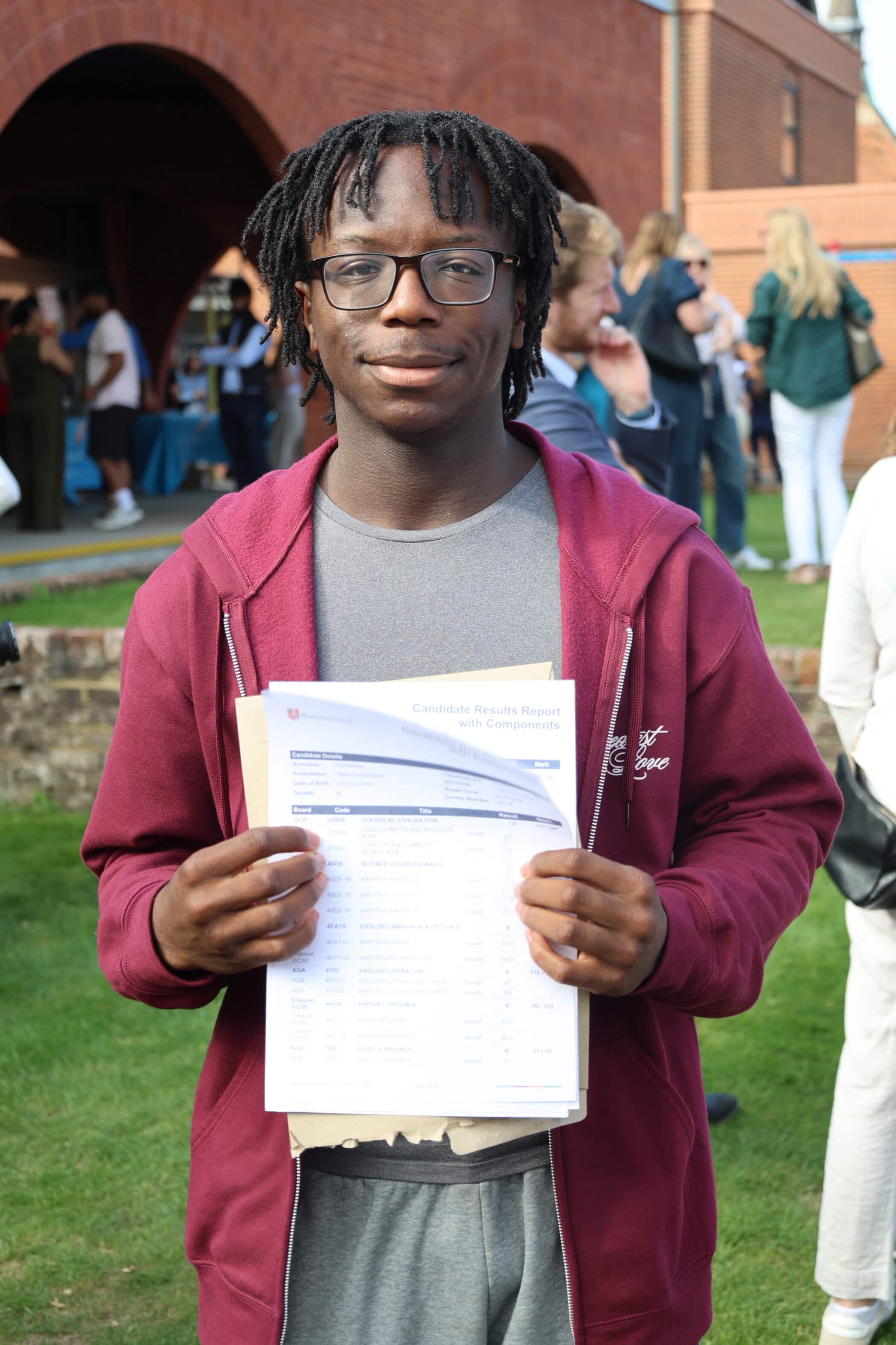 A young man wearing glasses and a maroon hoodie stands outside, holding his GCSE exam result papers and smiling. People and a brick building are visible in the background.