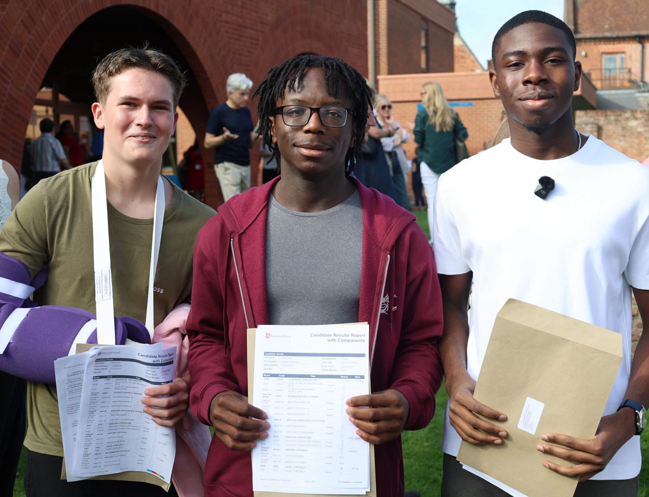 Three young men stand outdoors, smiling and holding their GCSE exam results and envelopes. One has his arm in a sling, and others stand in the background near a brick building on a sunny day.