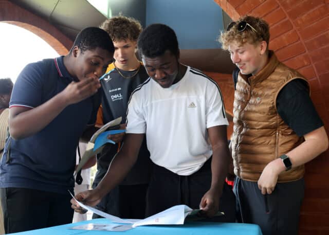 Four young men gather around a blue table, looking at GCSE papers with excited and surprised expressions. One wears a waistcoat, another sports a white shirt, and they are standing near a red brick wall.
