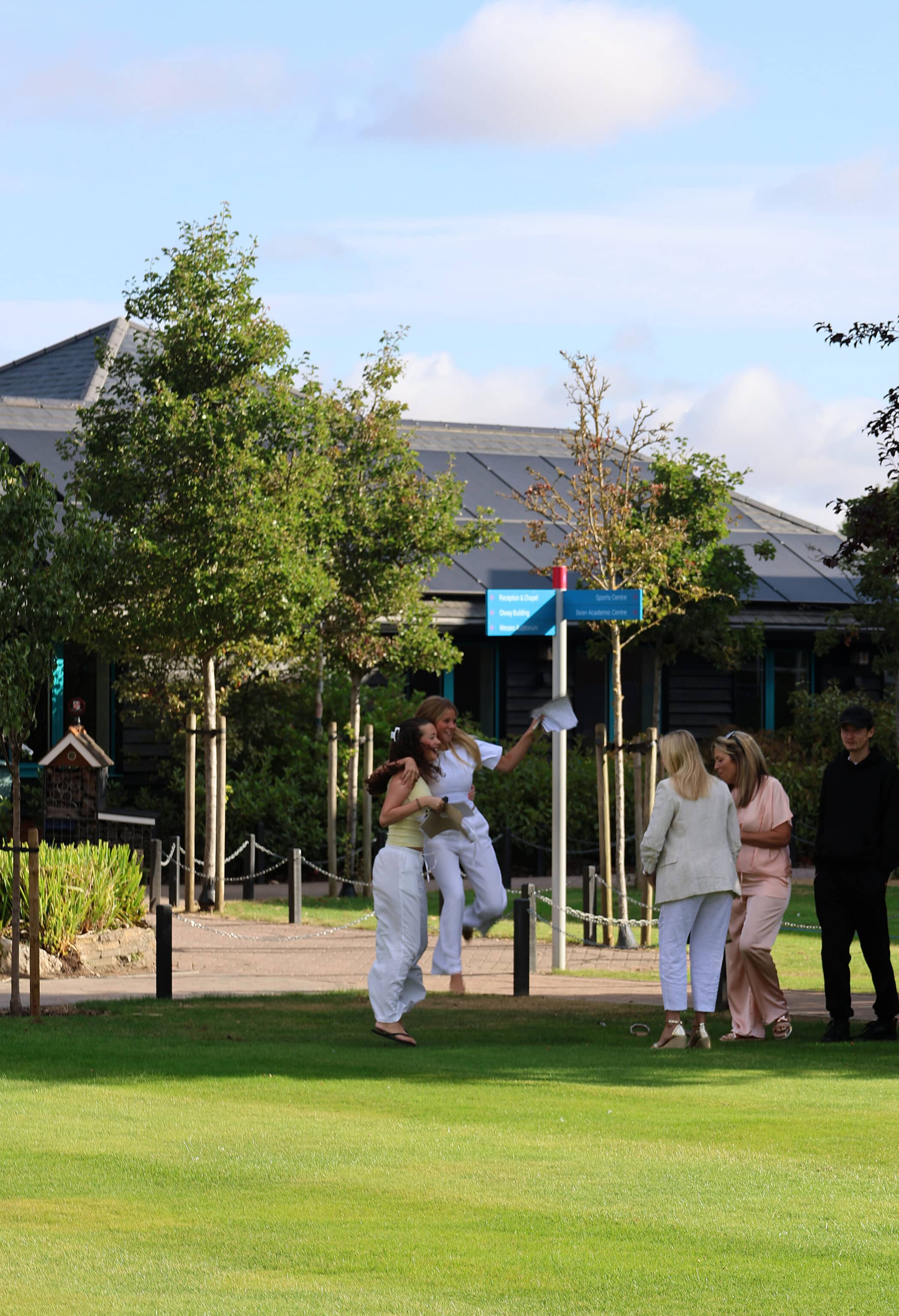 Two people in white jump and hug on a lawn, perhaps celebrating GCSE results, whilst three others in pastel clothes and one in black stand nearby. Trees and a building are in the background under a partly cloudy sky.
