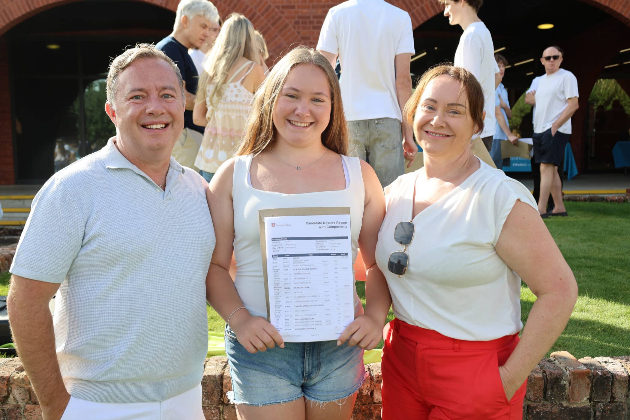 A smiling girl holds a GCSE results slip, standing outdoors between two adults. People are gathered in the sunny background near a brick building.