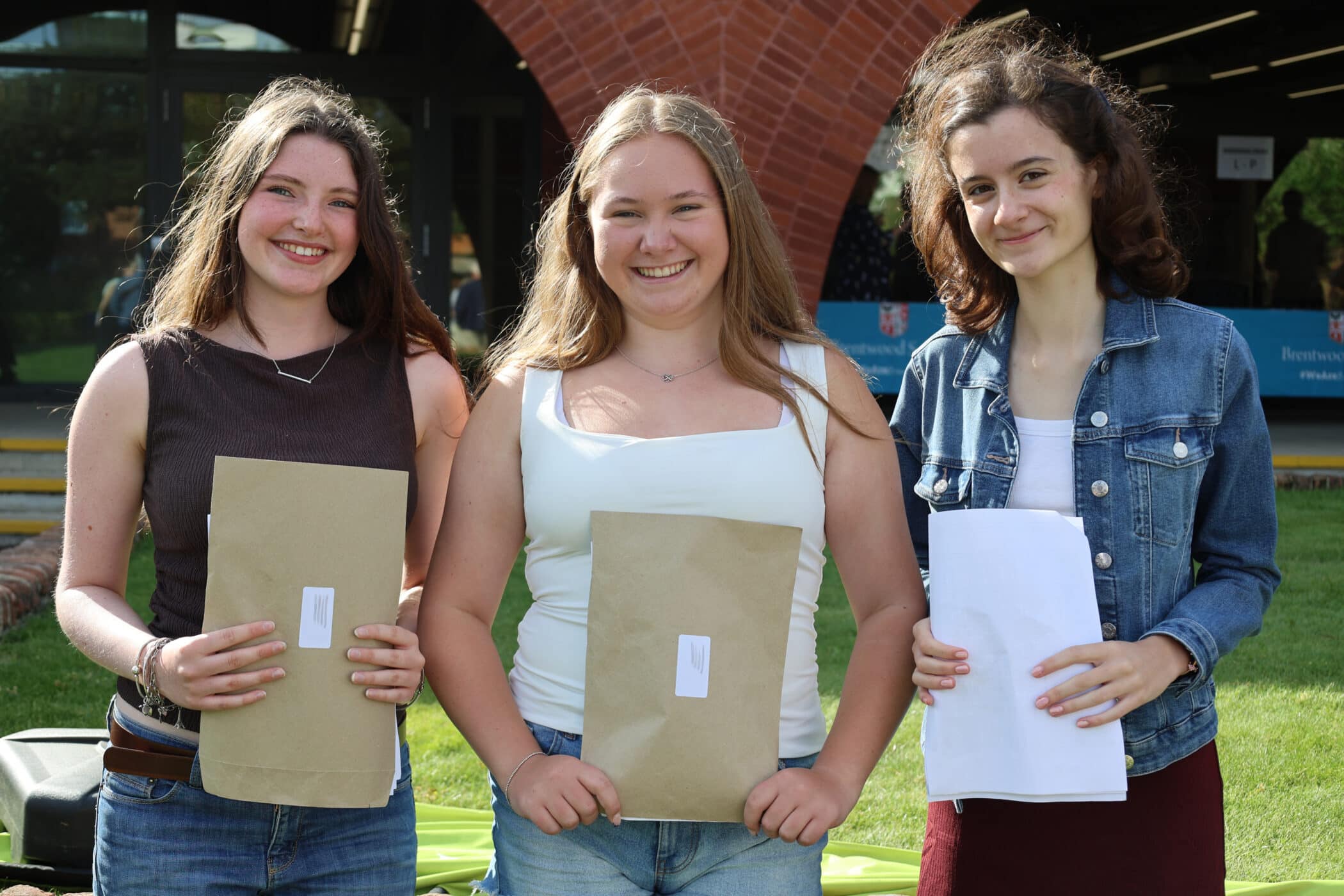 Three young women stand outside on a sunny day, smiling and holding large brown envelopes, possibly with GCSE results or important documents.
