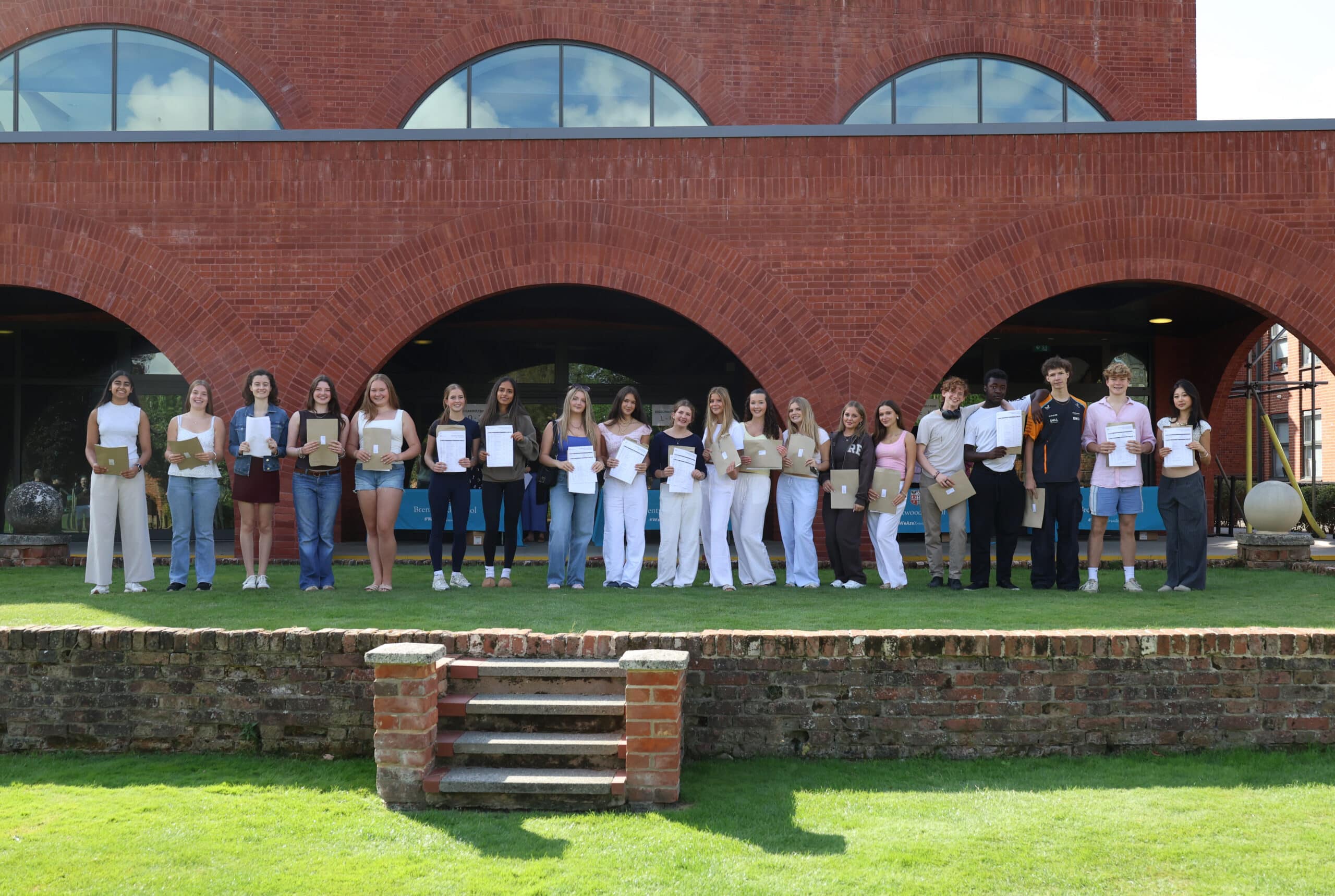 A group of young people stand in a queue outside a brick building, each proudly holding their GCSE certificates, smiling, and posing for a photo on a grassy lawn with a low brick wall in front of them.