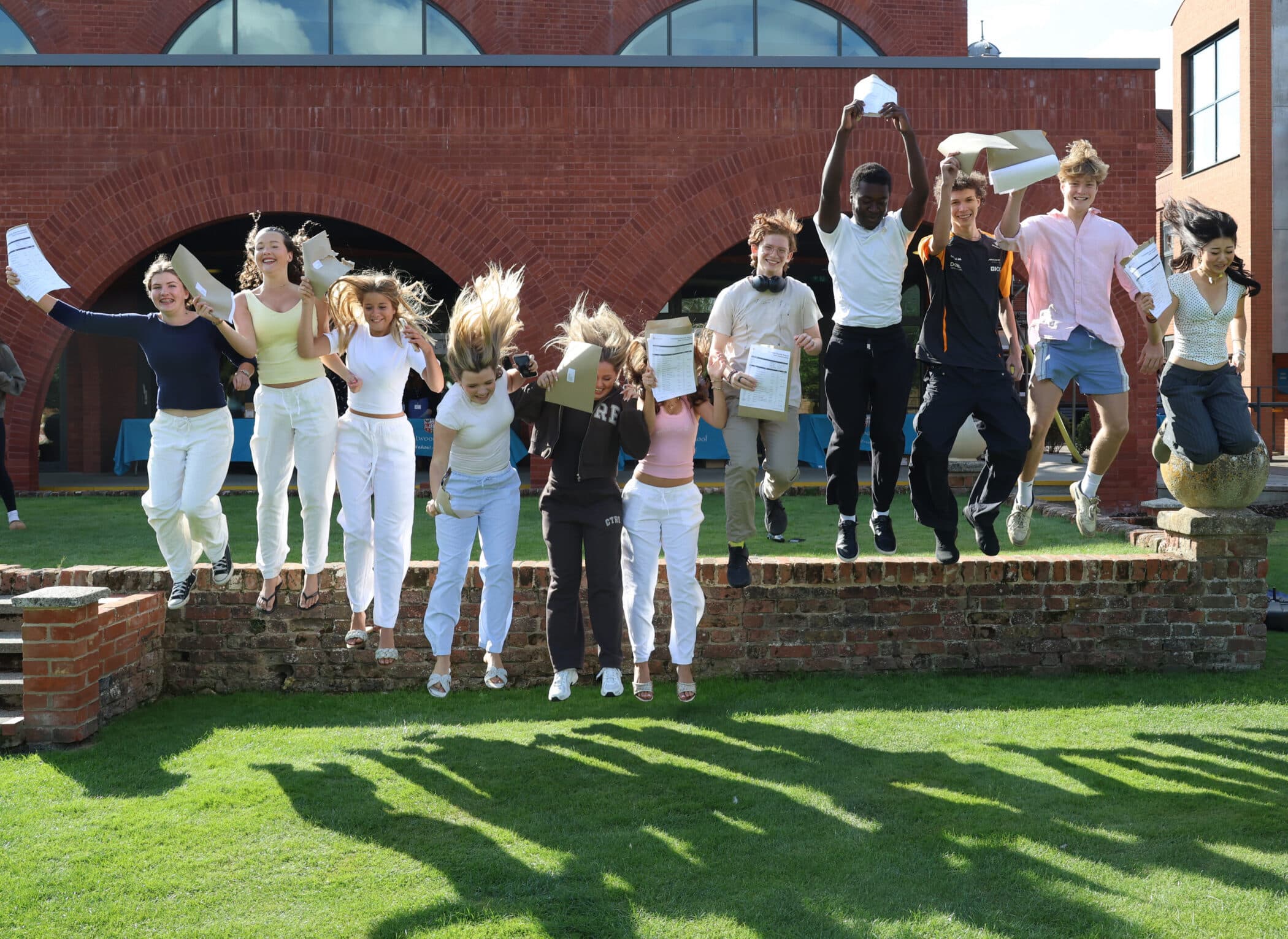 A group of students jump in the air, celebrating with their GCSE results papers in hand, outside a red brick building on a sunny day. Everyone appears happy and energetic.