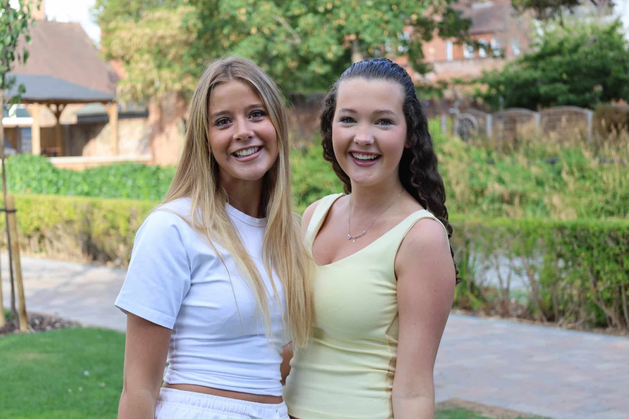 Two young women are smiling and standing close together outdoors on a sunny day, perhaps celebrating their recent GCSE results, with green foliage, trees, and buildings in the background.