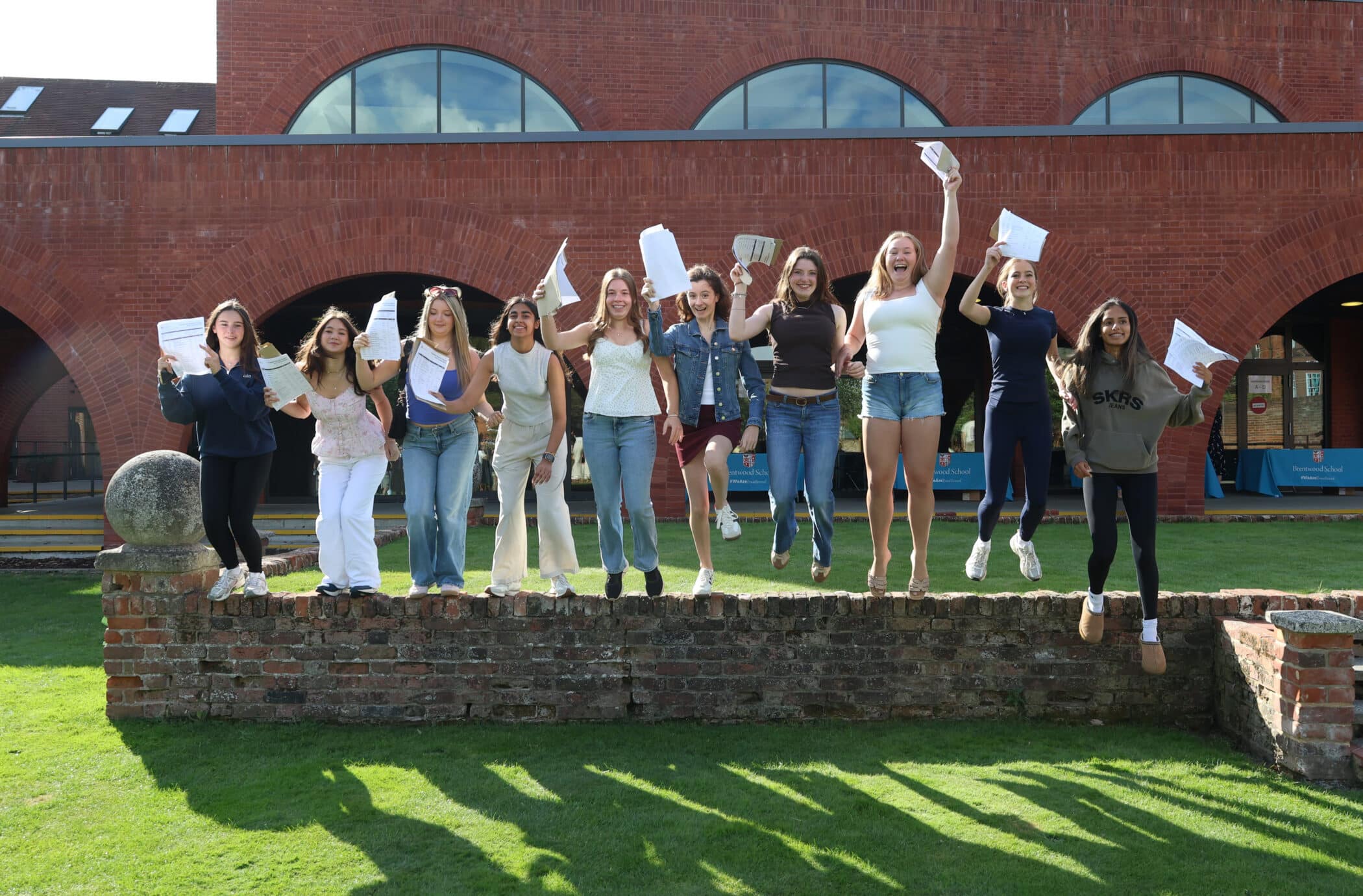 A group of young women stand on a low brick wall outside, jumping and holding GCSE papers in the air, celebrating together in front of a brick building with arched windows.