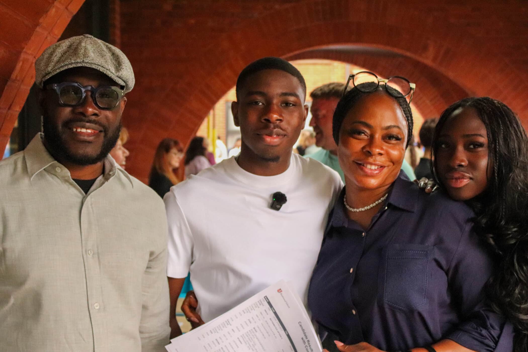 Four people stand together smiling, one proudly holding their GCSE results. They are outdoors with a brick arch in the background, dressed in casual to semi-formal clothing, and are celebrating an important achievement.
