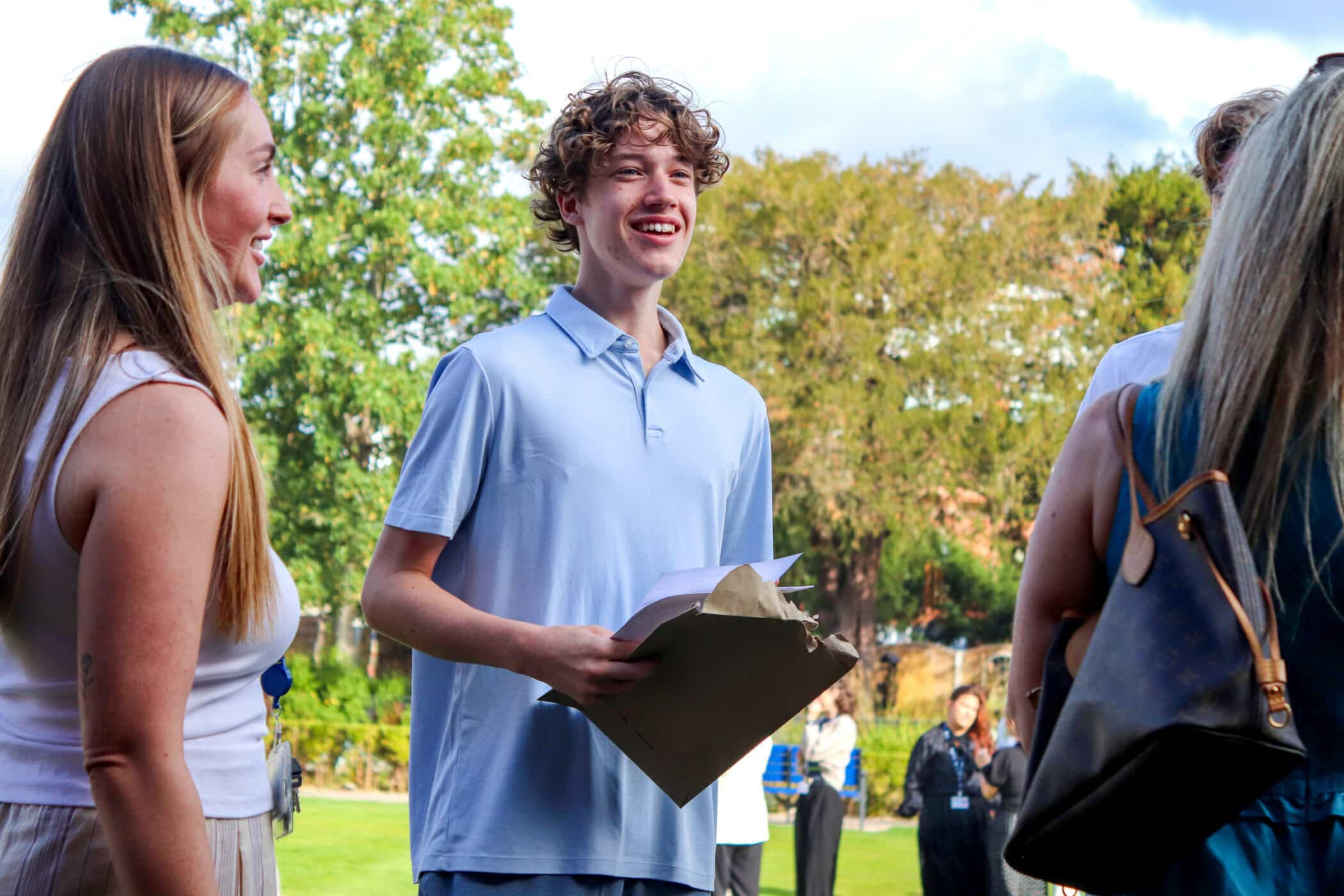 A smiling young person holding GCSE papers stands outdoors with a group of people, trees and grass in the background.