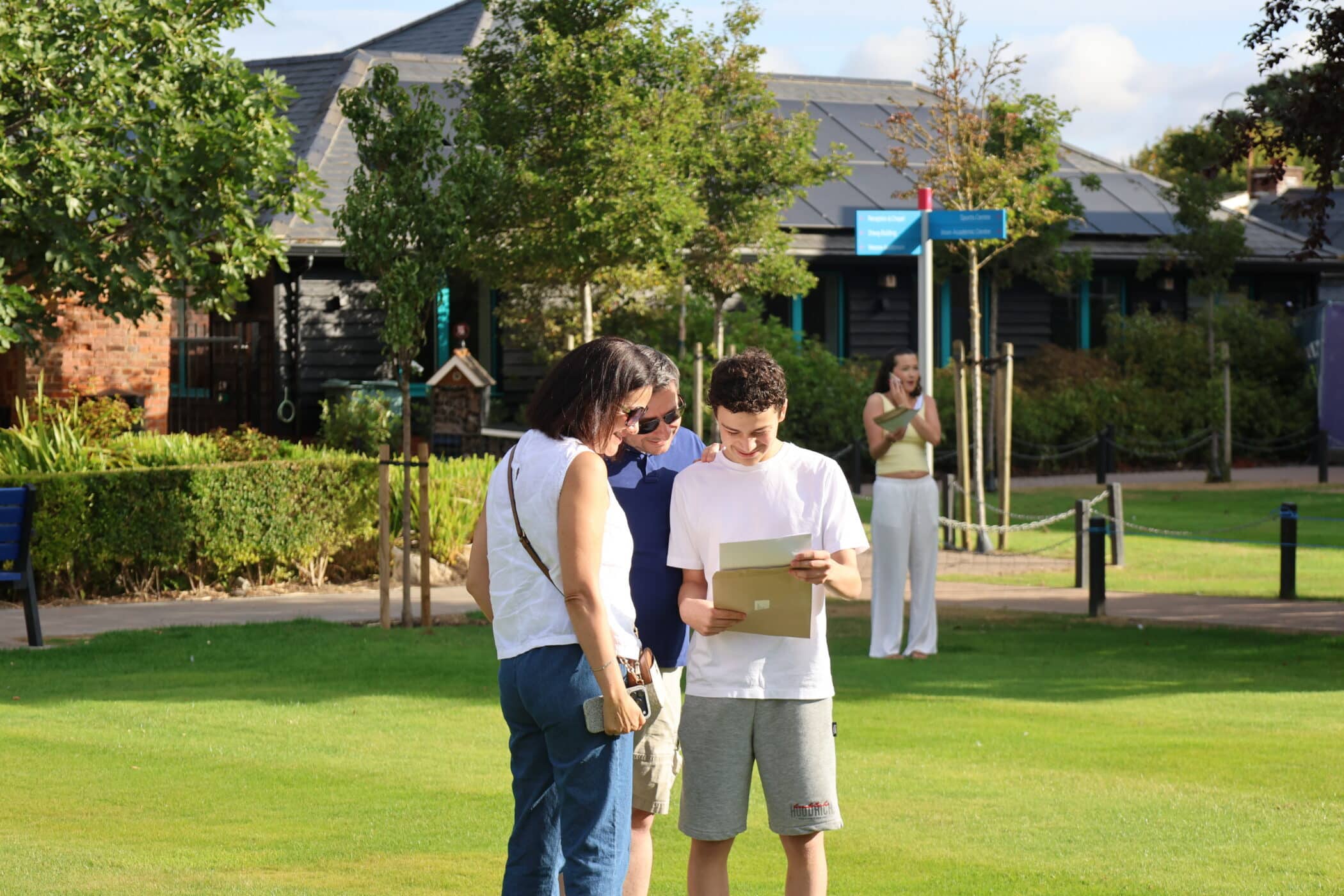 A teenage boy smiles whilst proudly holding his GCSE results with two adults beside him. A person in the background applauds, and they are all standing on a green lawn in front of a building on a sunny day.