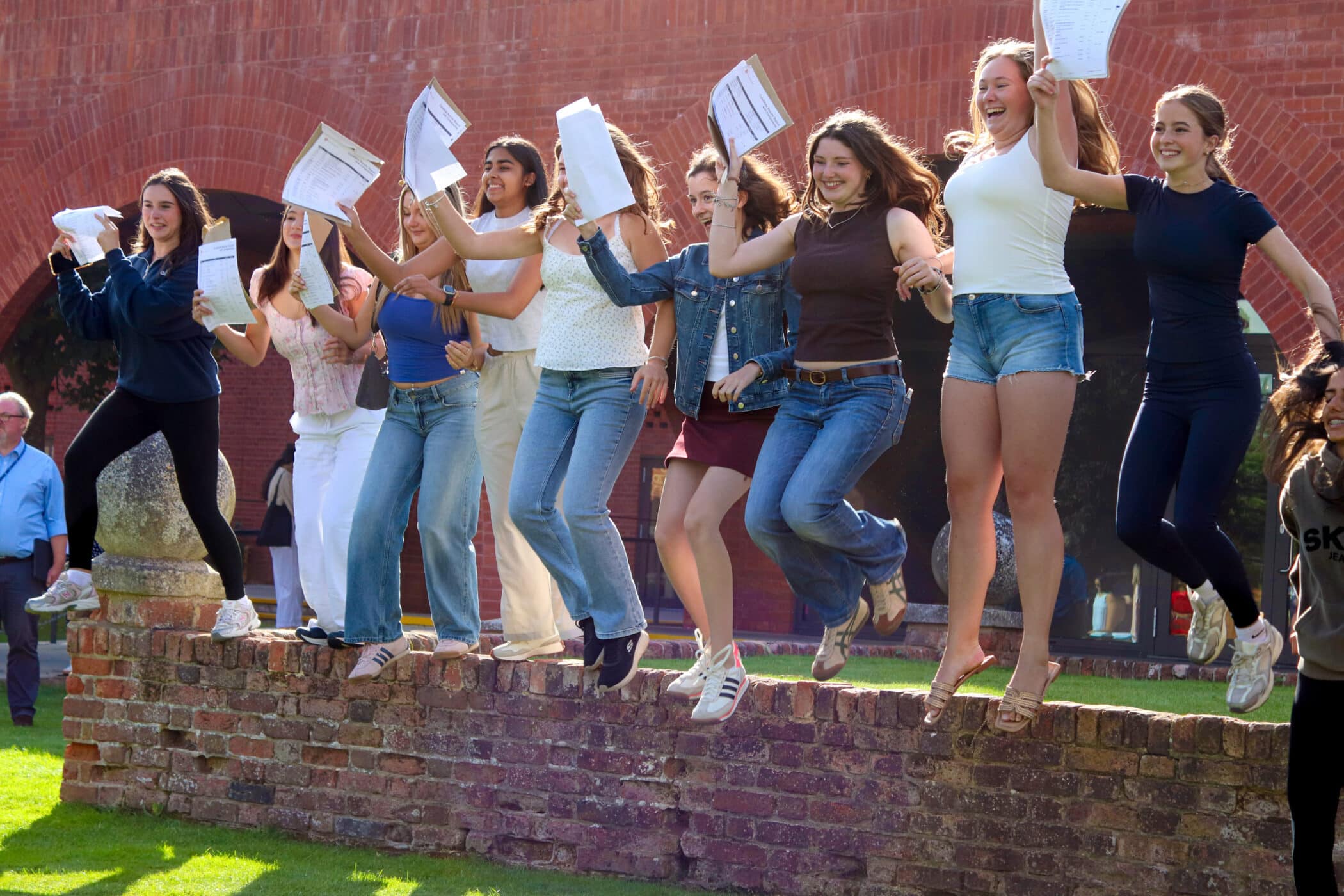 A group of young women joyfully jump off a low brick wall, holding their GCSE results and smiling, with a red brick building in the background on a sunny day.