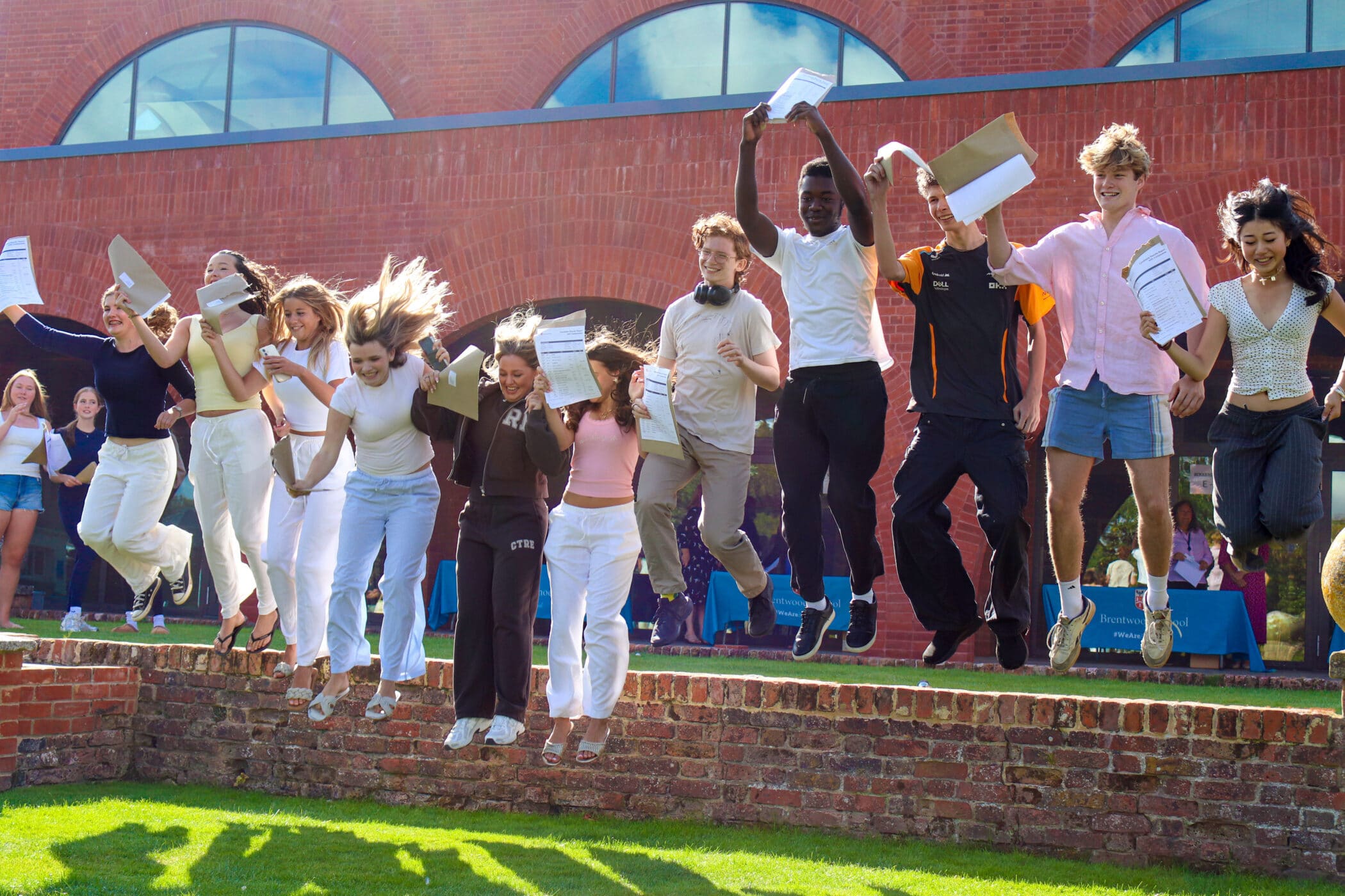 A group of students jump in the air, smiling and holding papers, in front of a red brick building. They appear to be celebrating their GCSE results on a sunny day.