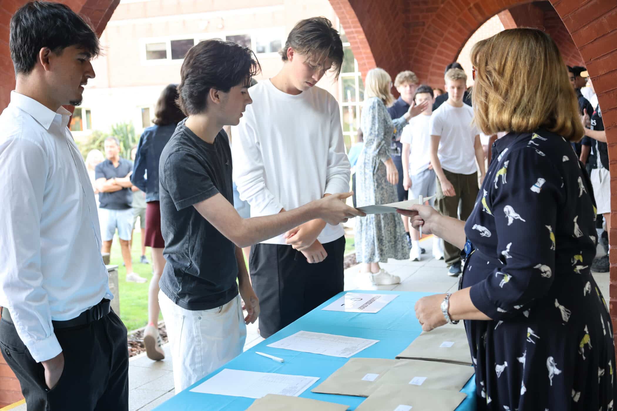 Students queue at a table to receive envelopes containing their GCSE results from a woman. Other people are gathered in the background under a brick archway on a bright day.