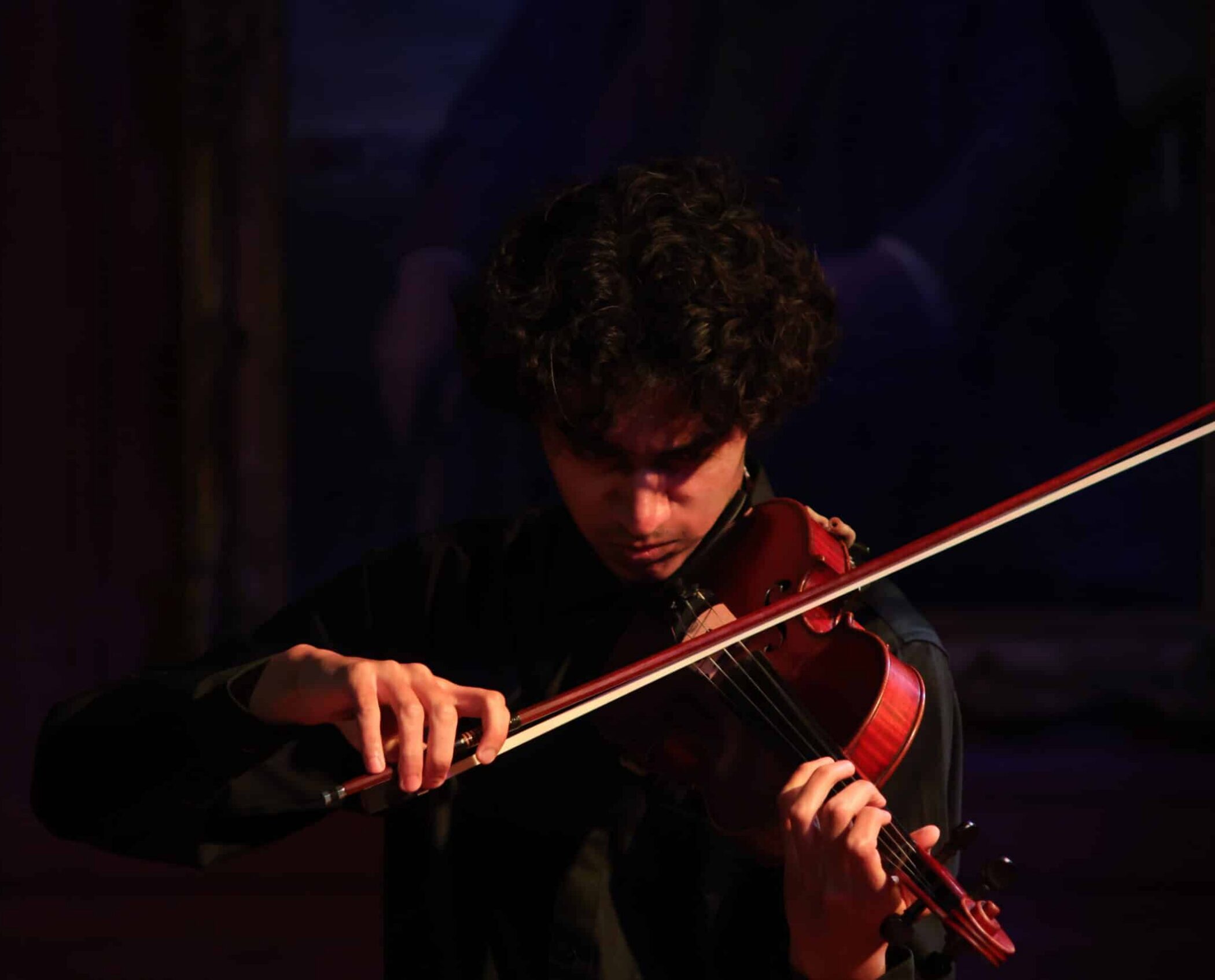 A person with curly hair, part of the Specialist Music Programme, plays a violin on stage under warm lighting, concentrating on their performance. The background is dark and slightly blurred, highlighting the musician and the instrument.