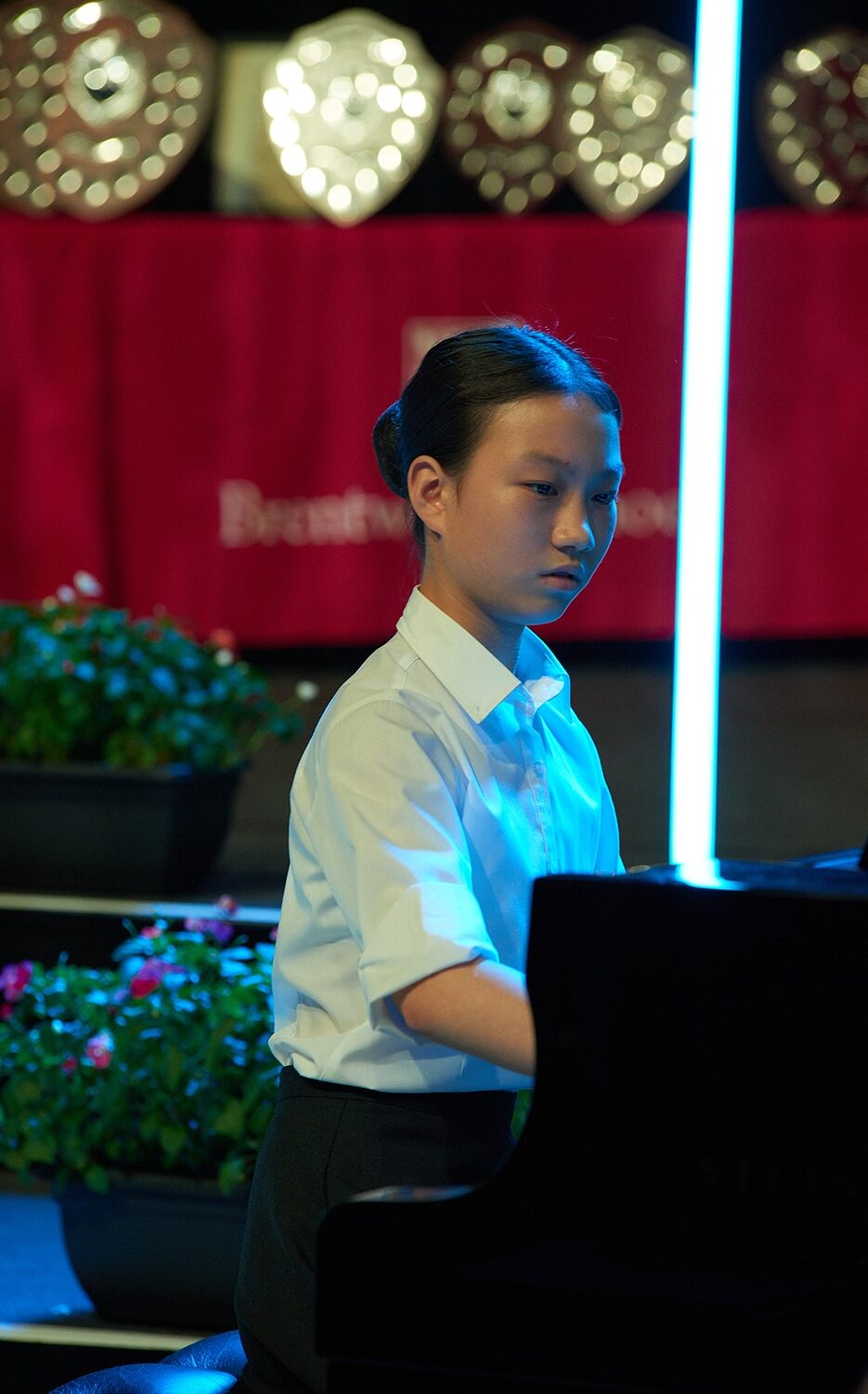 A young person in a white shirt, part of the Specialist Music Programme, plays piano on stage, lit by blue light. Trophies and red flowers are visible in the background.