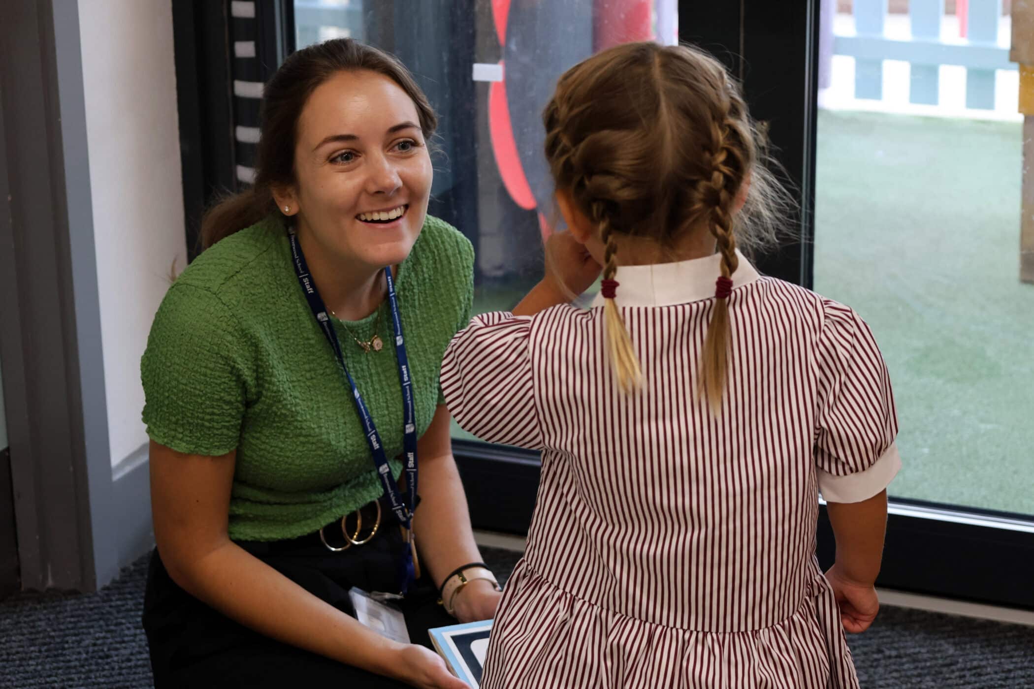 A woman in a green top smiles and talks to a young girl with plaited hair wearing a striped dress inside a Brentwood Preparatory School classroom near a glass door.