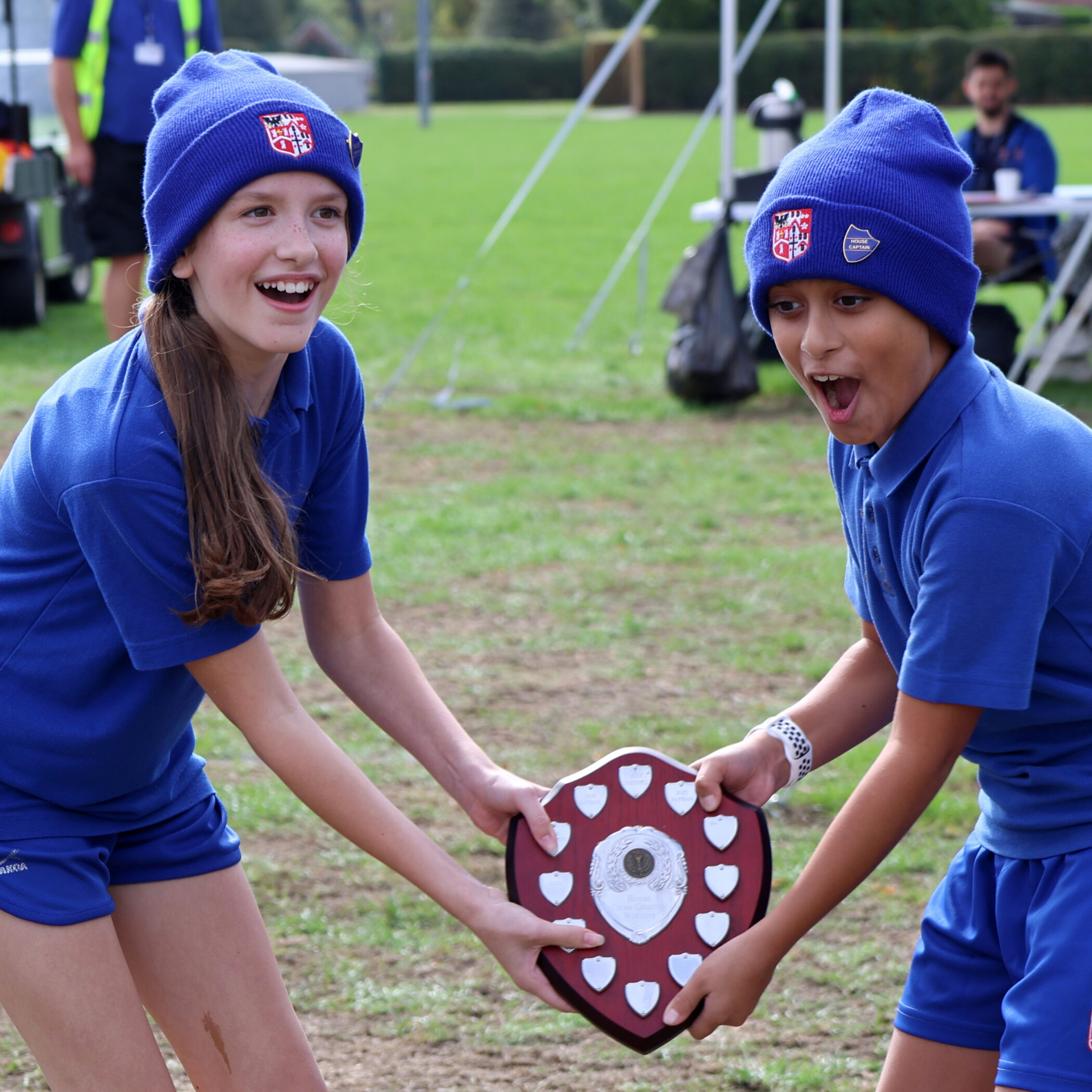 Two kids in blue uniforms and hats smile excitedly whilst holding a large shield-shaped trophy together on a grassy sports pitch.