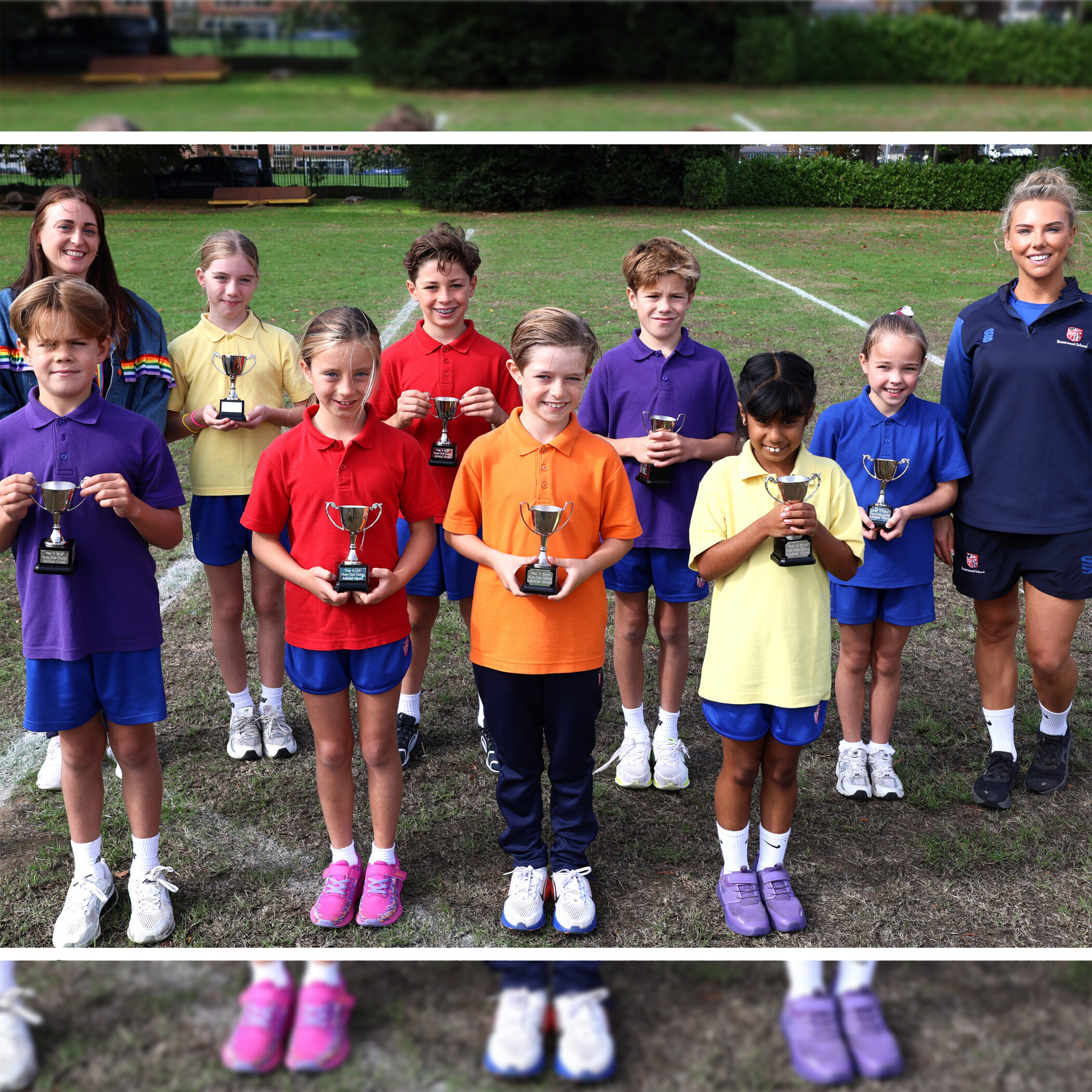 A group of children in colourful shirts hold trophies and stand outdoors with two adults on grass, smiling for a photo.