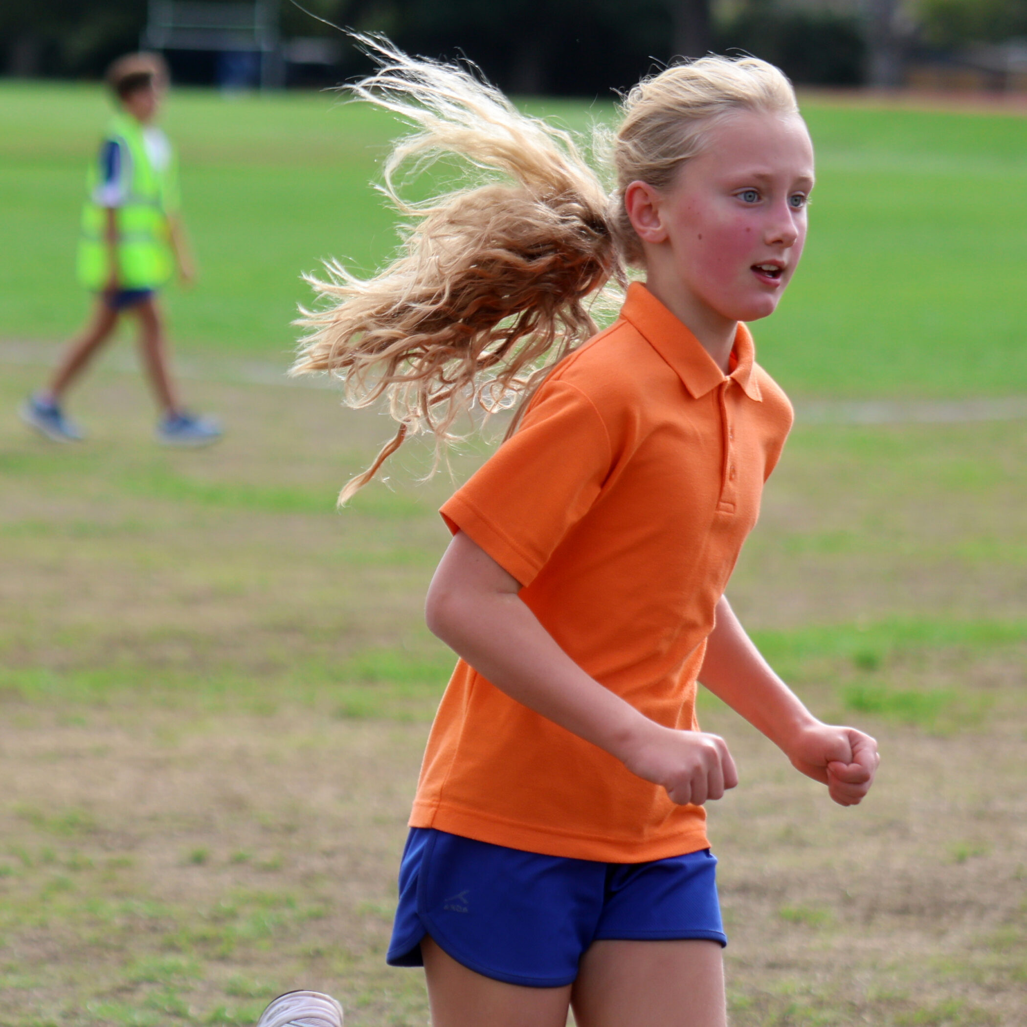 Young girl with blonde hair runs outdoors on grass, wearing an orange polo shirt and blue shorts. Another person in a reflective vest is blurred in the background. The setting appears to be a sports pitch.