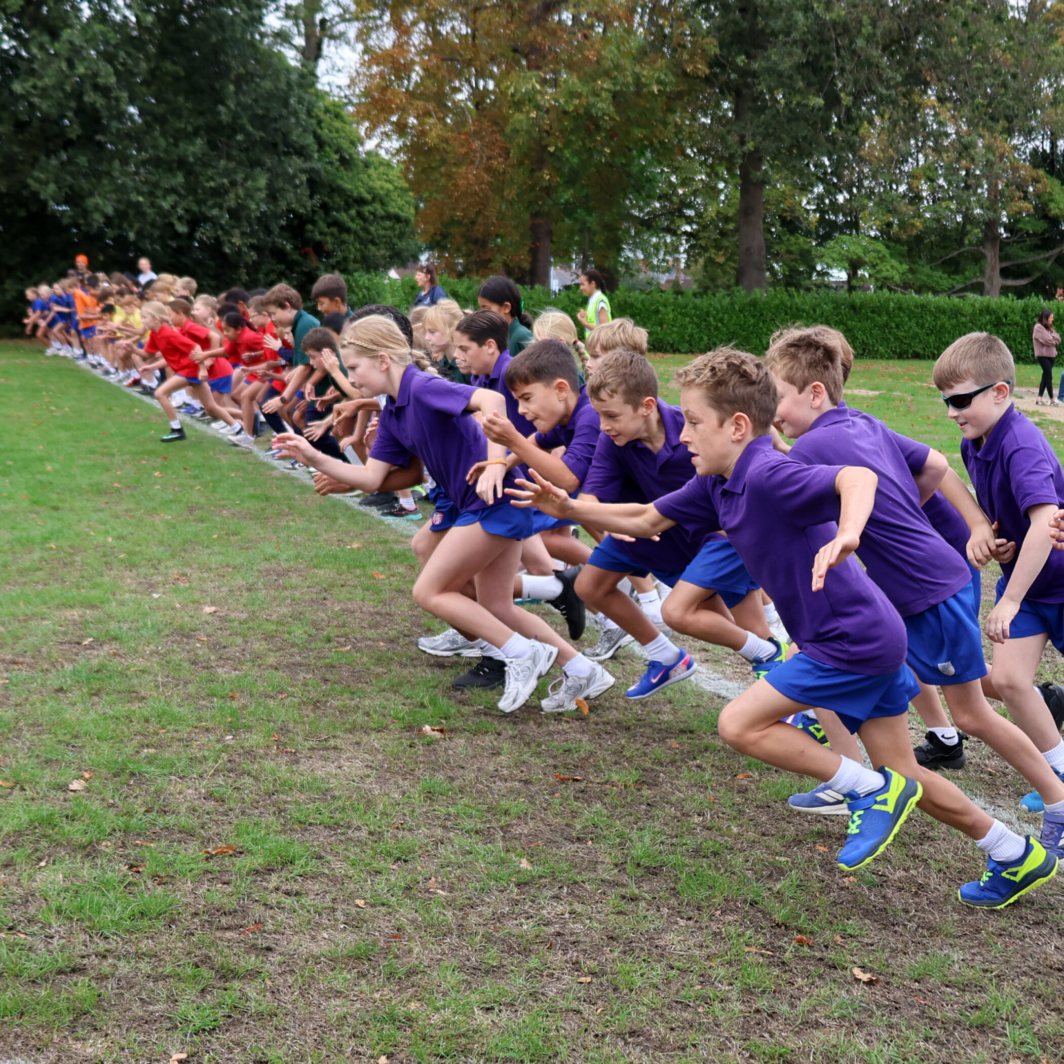 A large group of children in coloured shirts are starting a race on a grassy field, energetically sprinting forward from the starting line outdoors.
