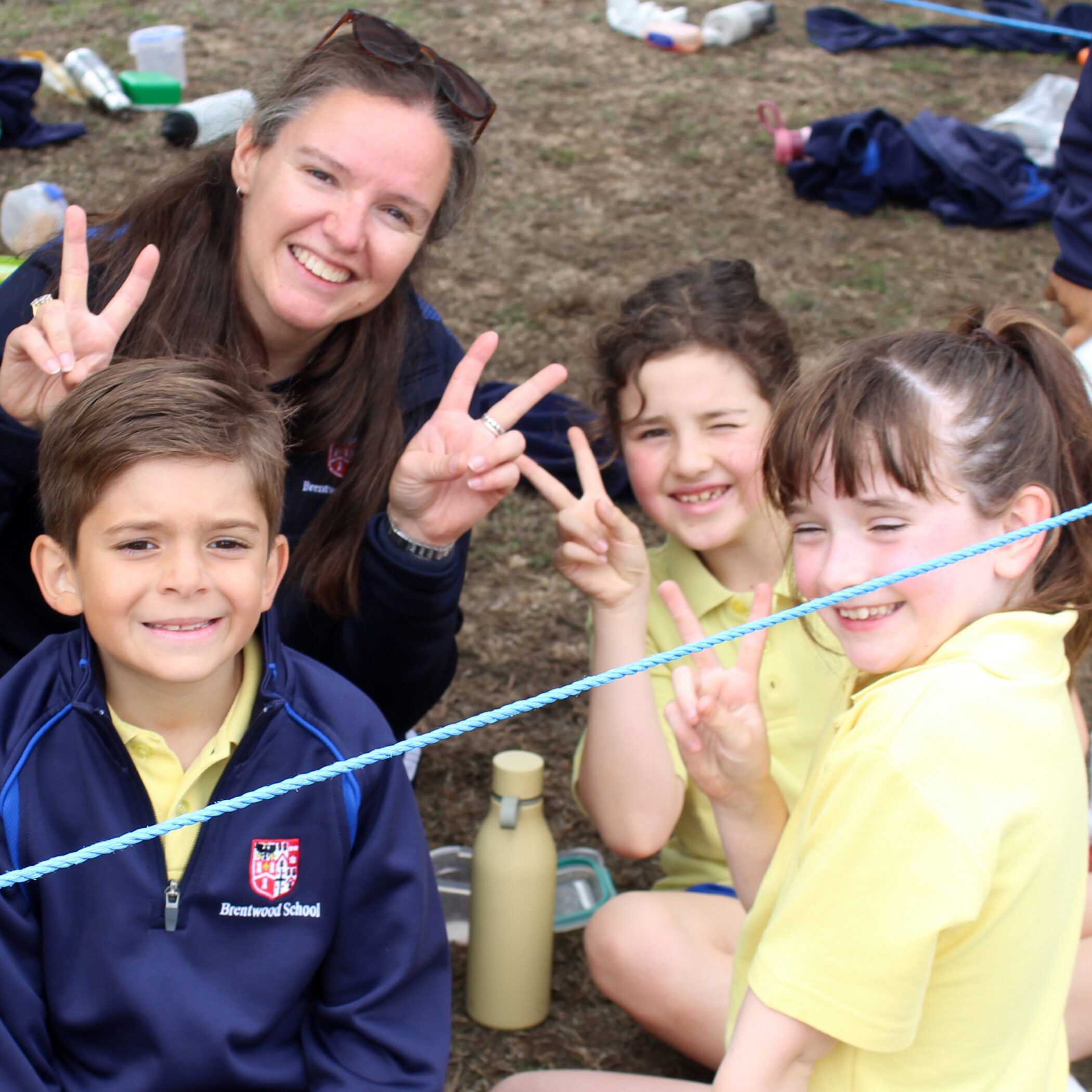 A woman and three children sit outside, smiling and making peace signs with their fingers. They are wearing school uniforms and a water bottle is visible near them. A blue rope runs across the foreground.