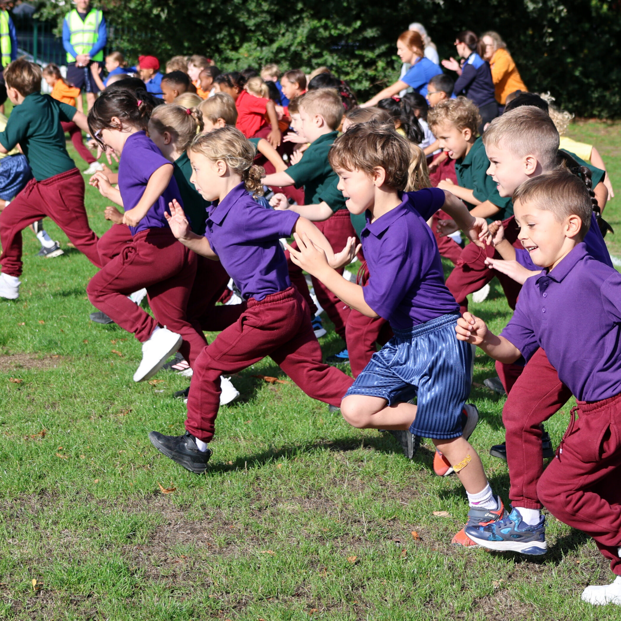 A group of children in colourful uniforms are running on a grassy field, participating in an outdoor race or event on a sunny day.