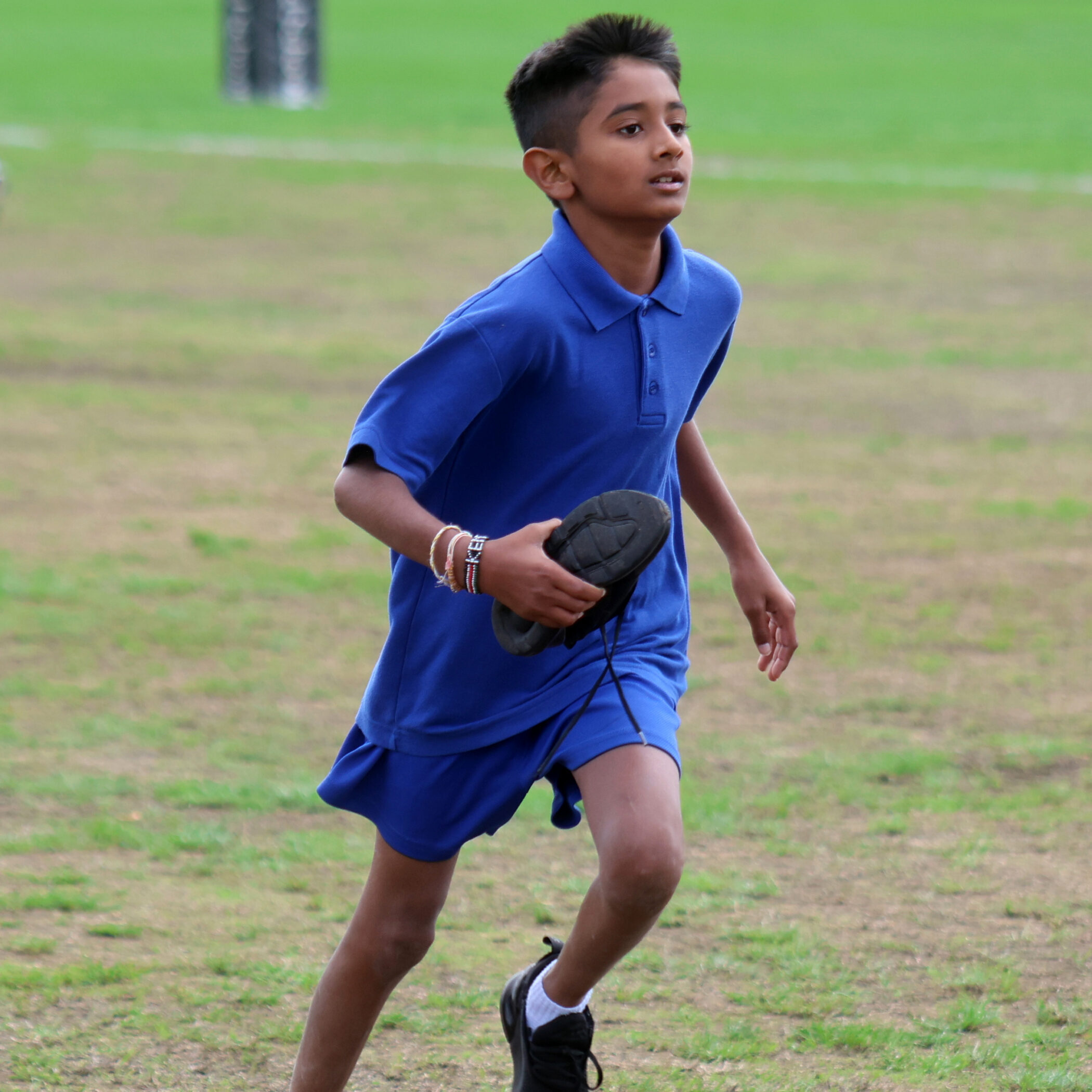A boy in a blue shirt and shorts runs on grass, holding a black shoe in his right hand. The background shows a green field.