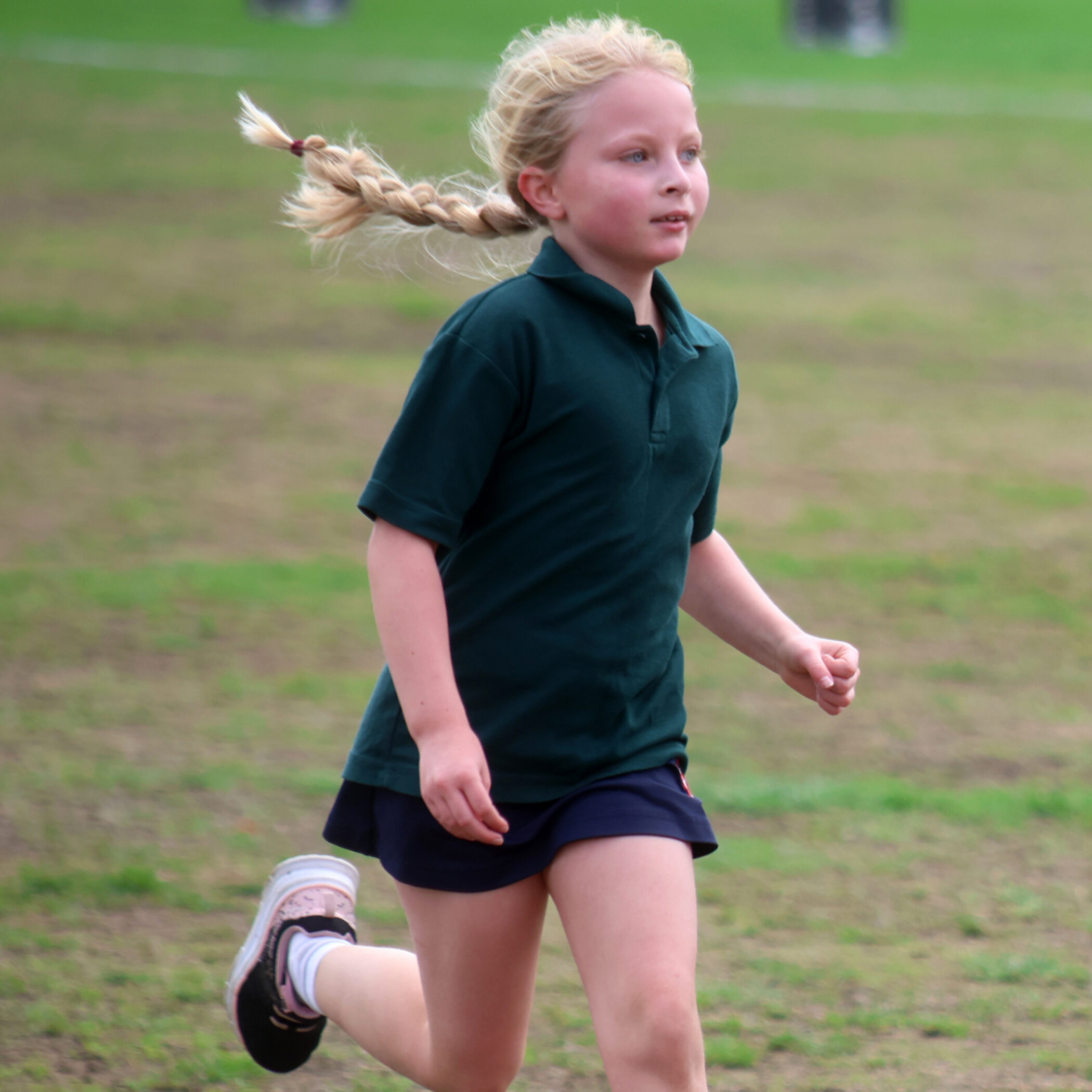 A young girl with a plaited ponytail runs outdoors on a grassy field, wearing a dark green polo shirt, navy shorts, and trainers.