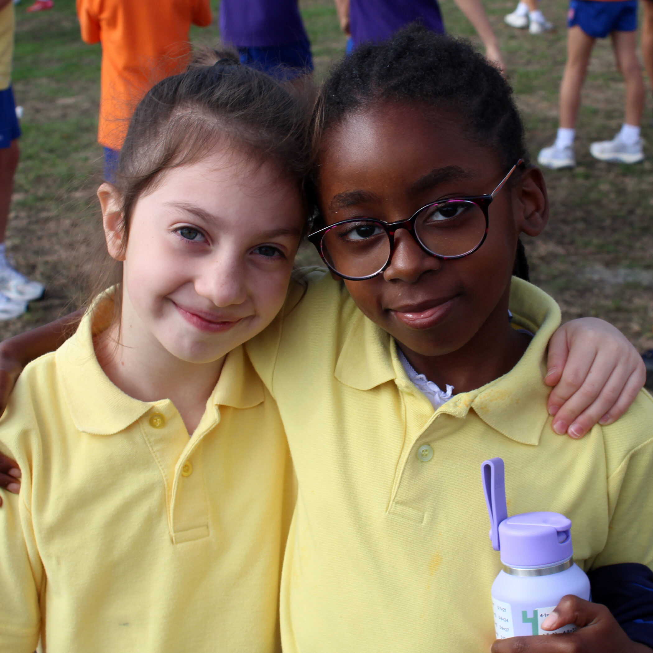Two children wearing yellow shirts stand close together, smiling at the camera with arms round each other. One child holds a water bottle; other children are visible in the background.
